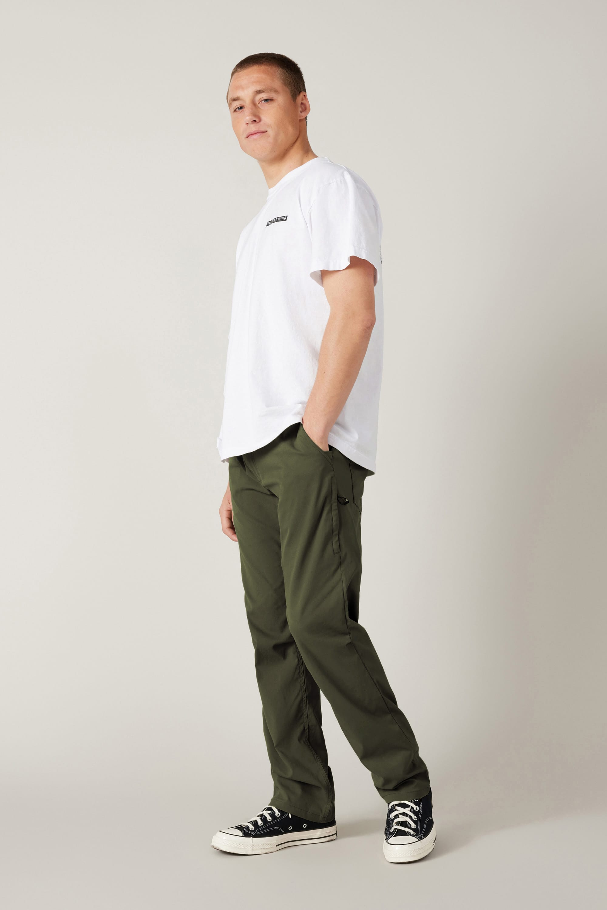 A person stands in a relaxed pose wearing a white T-shirt, 686 Men's Everywhere® Pant - Relaxed Fit in olive green by 686, and black-and-white sneakers against a plain light gray background.