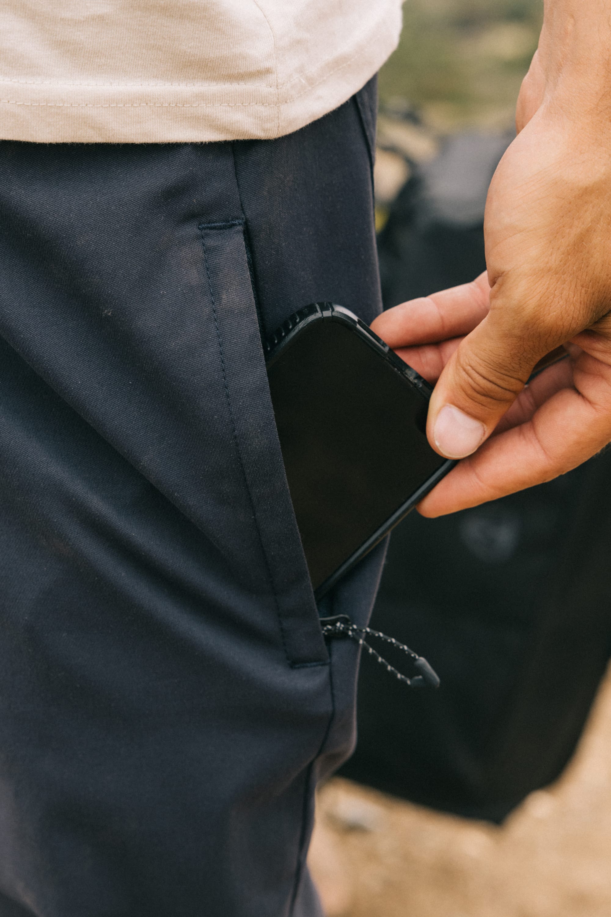 A person in 686 Men's Everywhere® Pant - Relaxed Fit puts a black smartphone into their front pocket with their right hand, while the background is slightly blurred.