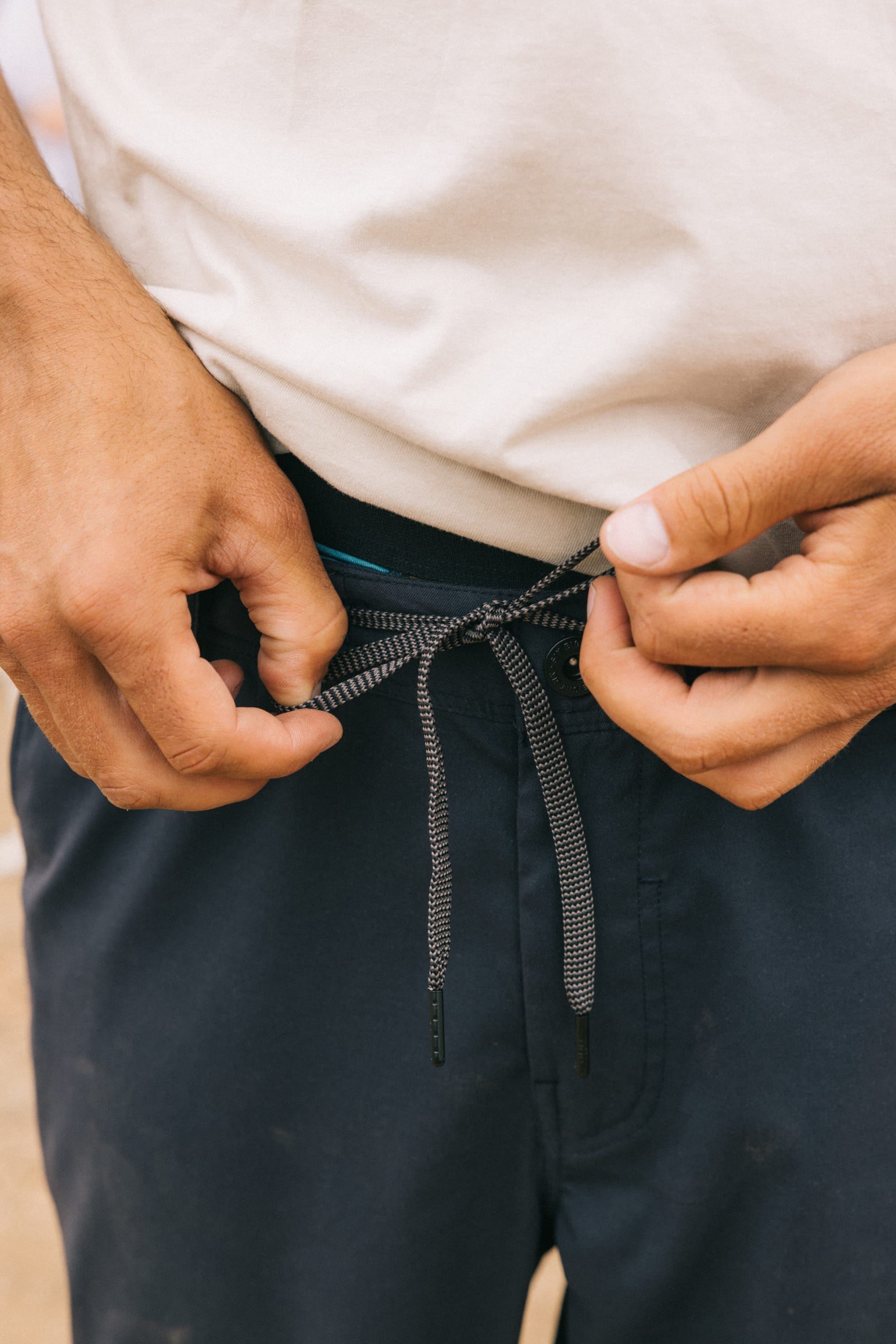 A person ties the black-and-white drawstring of their 686 Men's Everywhere® Pant - Relaxed Fit in dark blue while wearing a white shirt. Only their hands, torso, and part of their clothing are visible. Brand: 686.