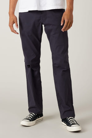 A person is shown from the waist down, wearing 686 Men's Everywhere® Pant - Relaxed Fit in dark navy, paired with a white t-shirt and black low-top sneakers, standing against a plain light background.