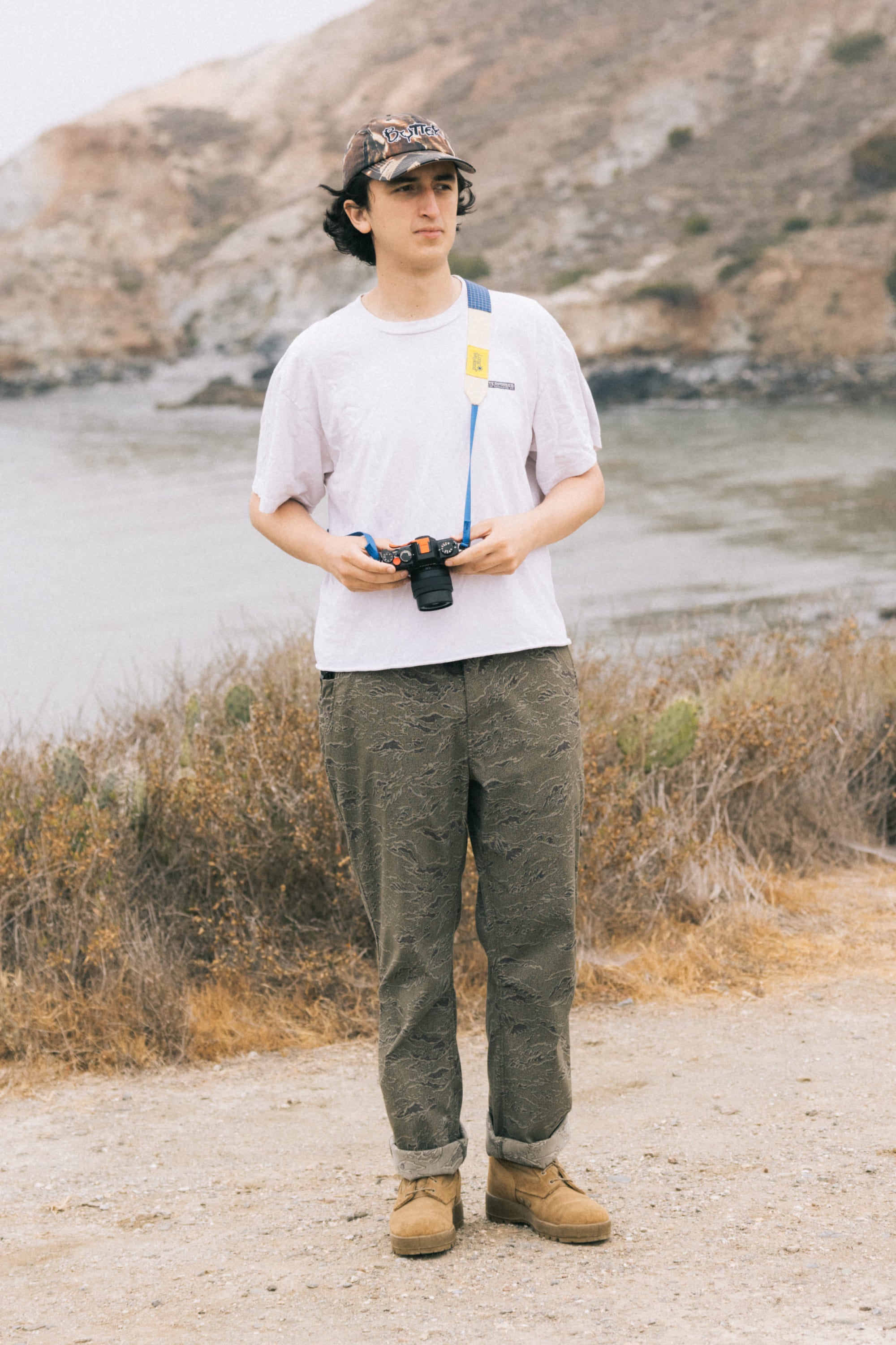 A person in a white t-shirt, 686 Men's Everywhere® Pant - Relaxed Fit by 686, and tan shoes stands on a dirt path near dry bushes with a camera. A rocky hillside and water are visible in the background.