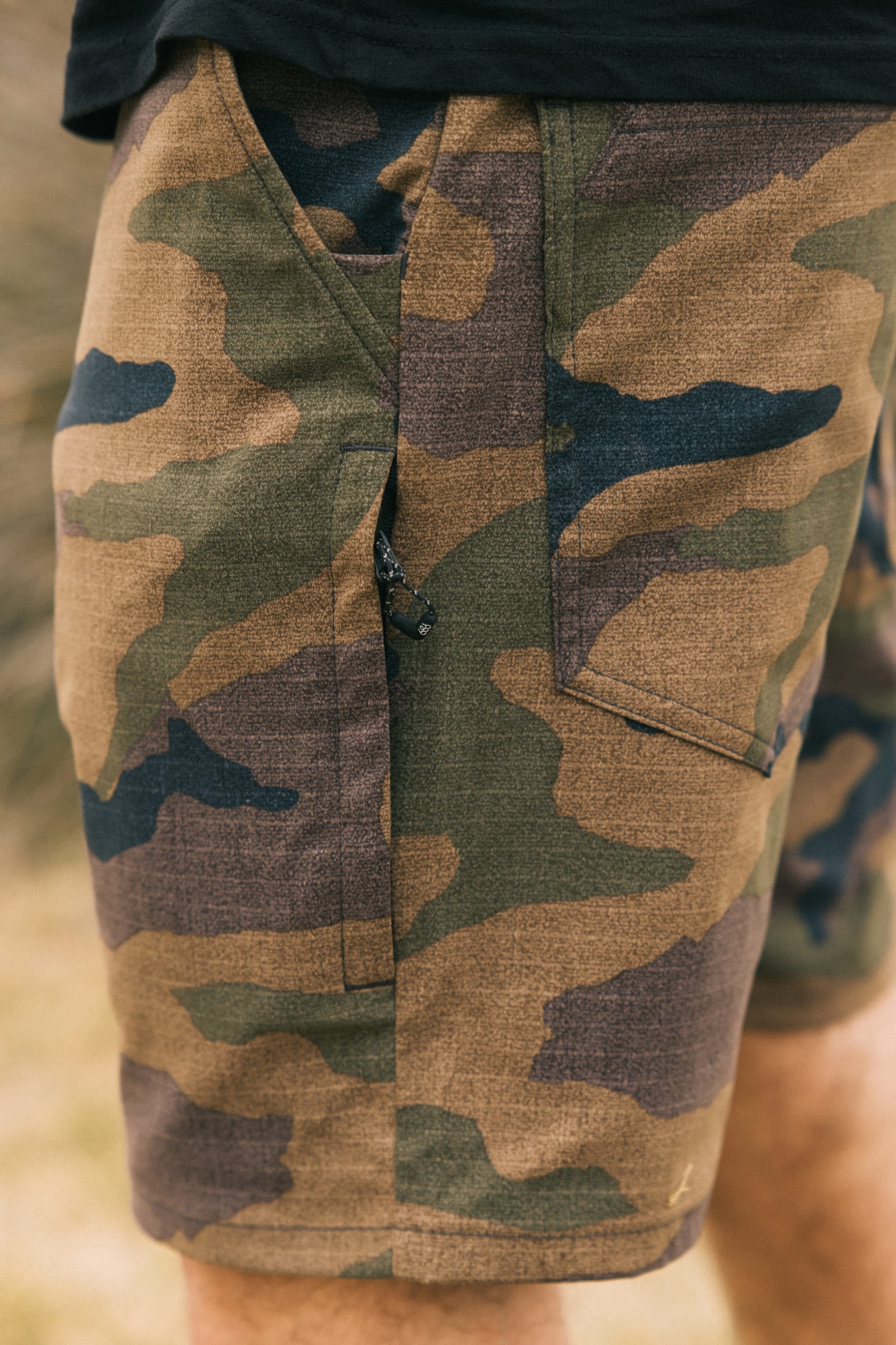 Close-up of a person wearing 686 Men's Everywhere® Hybrid Short by 686, featuring green and brown camouflage, pocket and zipper detail, breathable fabric, outdoors.