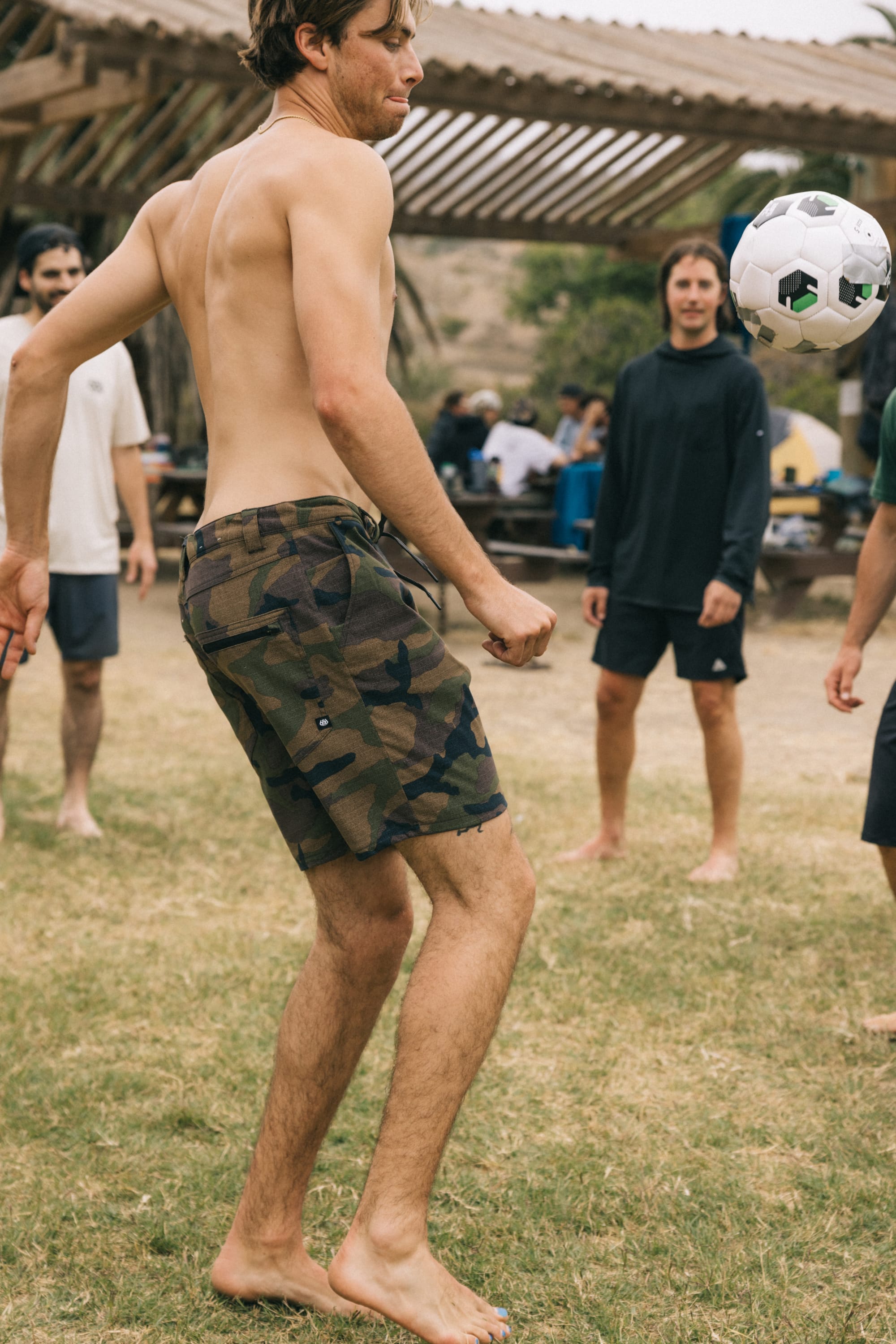 A barefoot man wearing 686 Men's Everywhere® Hybrid Short skillfully balances a soccer ball on his thigh while playing with friends outdoors, as others watch from the grassy area nearby.