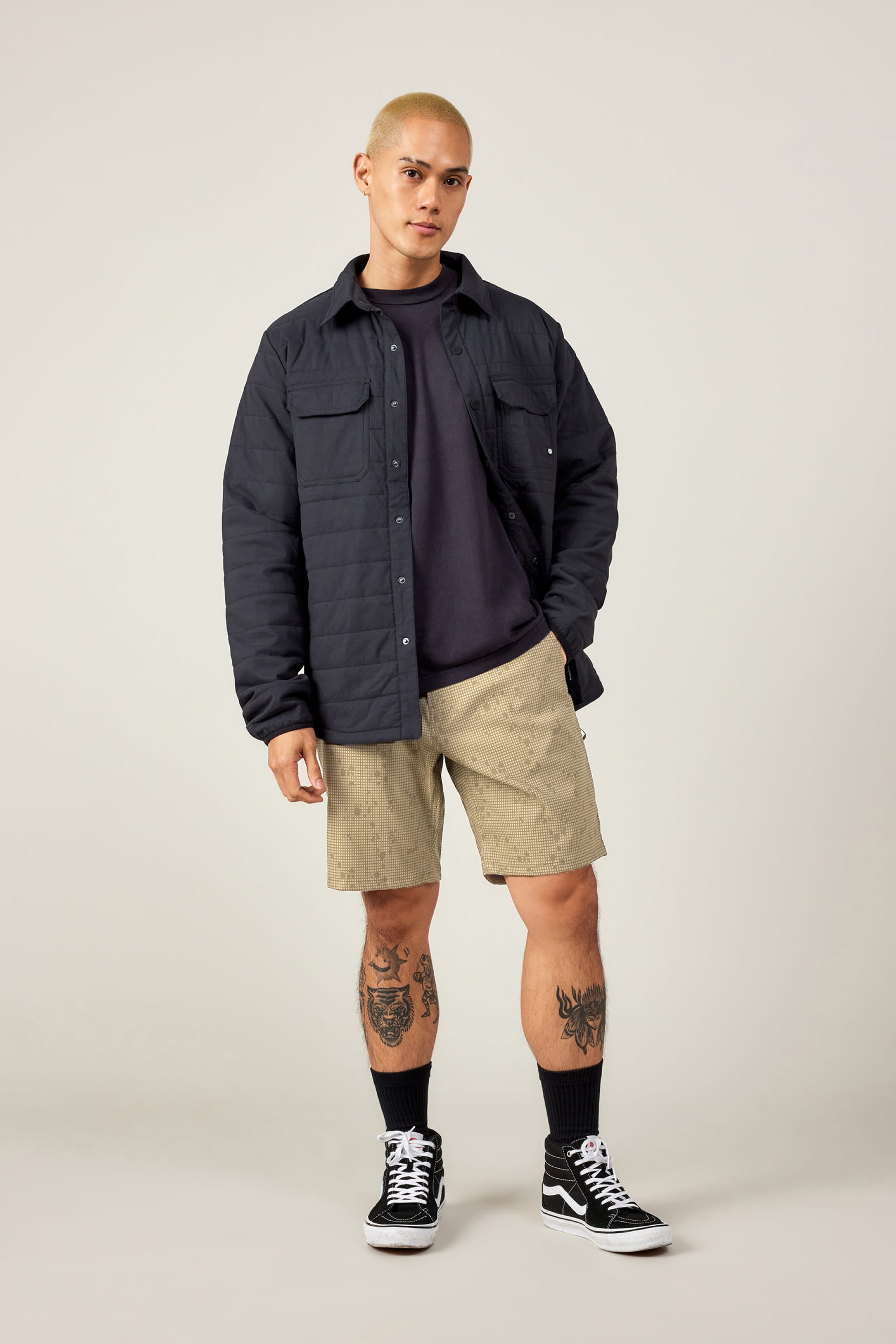 A person with short blond hair wears a black quilted jacket, dark T-shirt, 686 Men's Everywhere® Hybrid Short by 686 with patterns, black socks, and sneakers. Tattoos cover both legs as they stand against a plain light background.