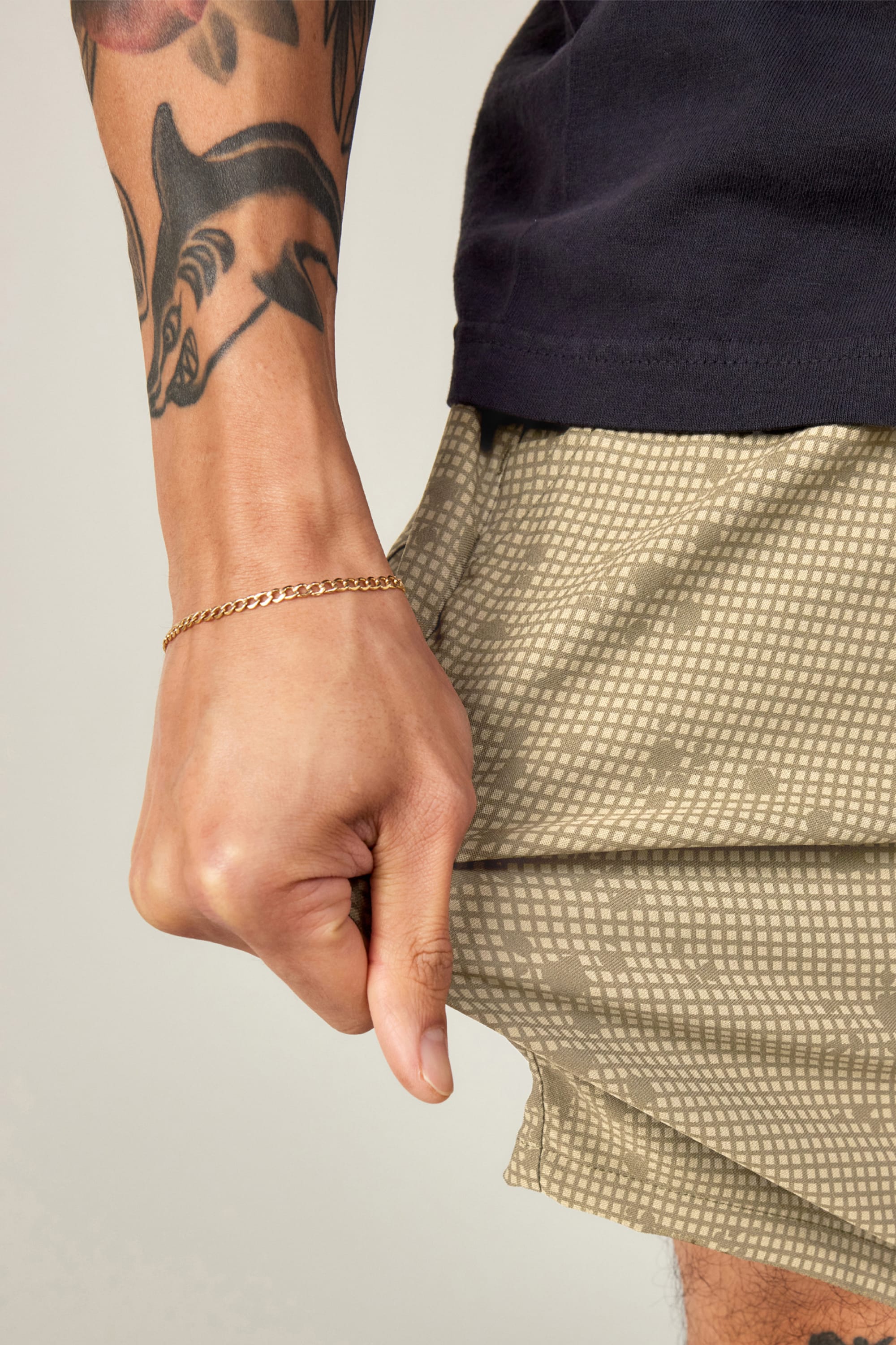 A close-up of a tattooed arm wearing a thin gold chain bracelet, holding the hem of 686 Men's Everywhere® Hybrid Short from 686—breathable, stain-resistant shorts—paired with a dark shirt.