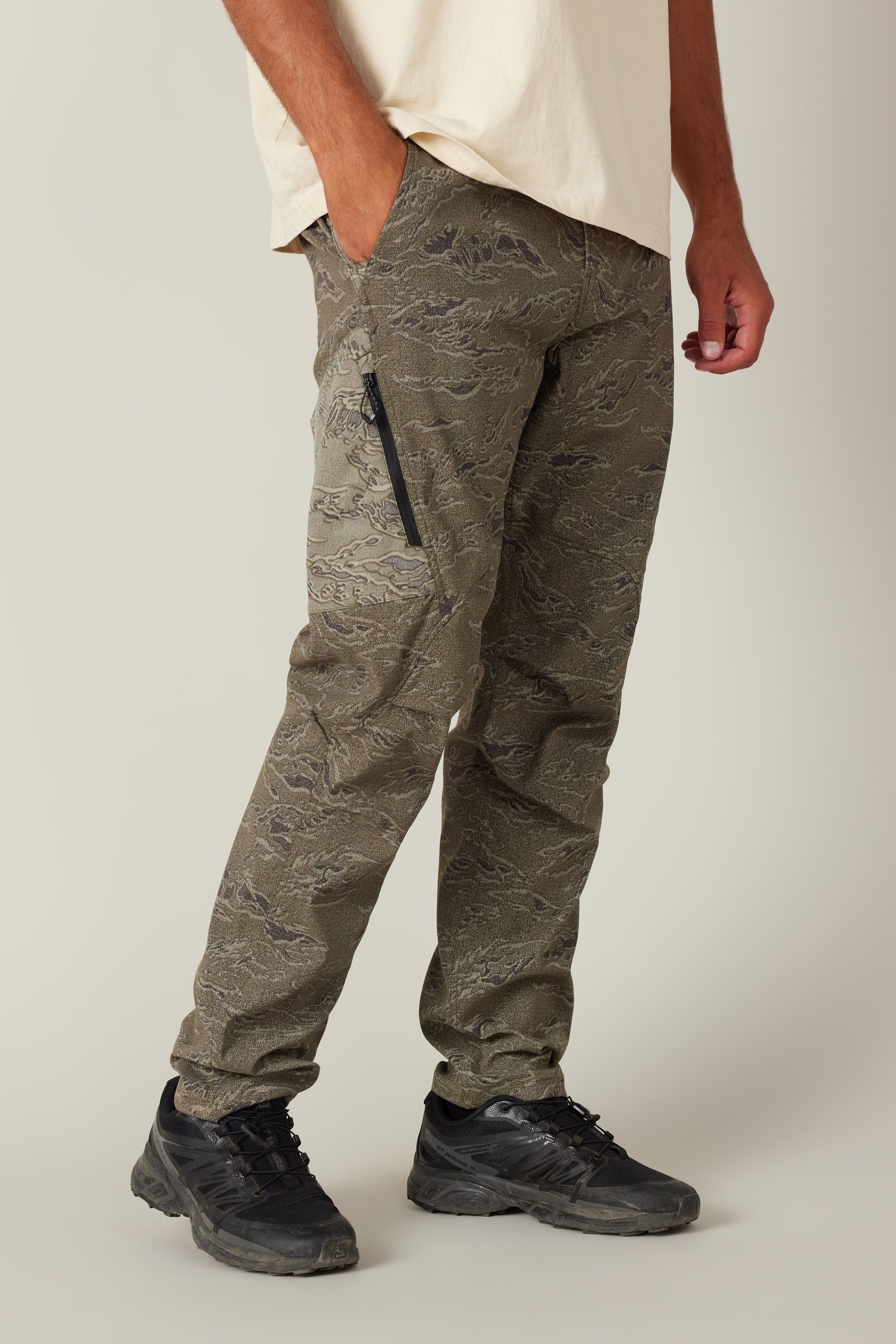 Wearing the 686 Men's Anything Cargo Pant - Slim Fit by 686, featuring a 13-pocket design and zippered side pocket, and a cream tee with black sneakers, a person stands with one hand in their pocket against a neutral background.