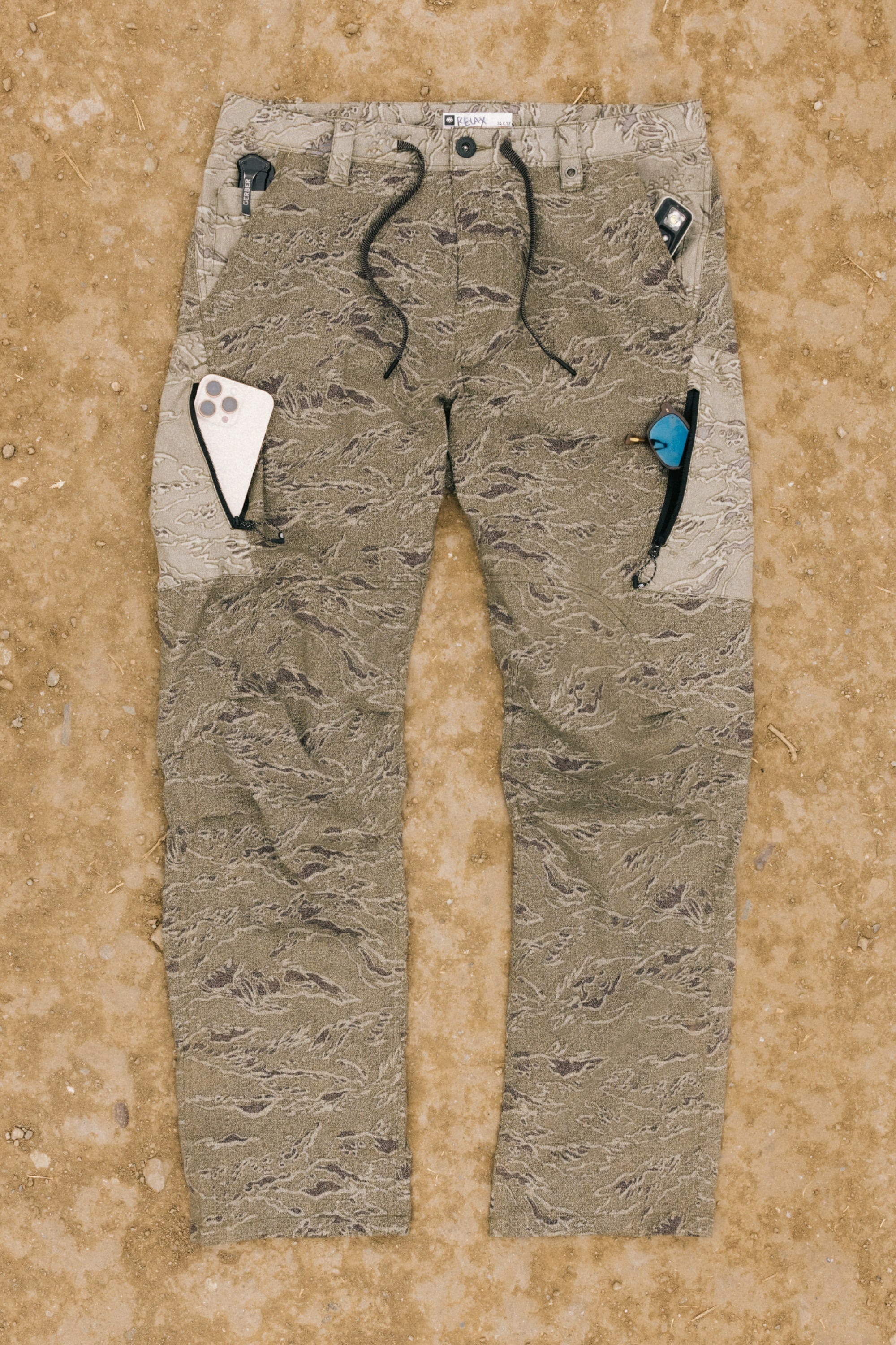 A pair of 686 Men's Anything Cargo Pant - Slim Fit with 13 pockets and camouflage pattern lies flat on dirt. Drawstrings, visible seams, and multitools in the pockets highlight its technical, adventure-ready design.