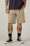 A person wears a dark t-shirt, 686 Men's Everywhere® Hybrid Short - Relaxed Fit in beige check, black socks, and black Converse sneakers. Their tattooed arms and legs stand out against a plain background.