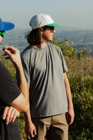 Two young men outdoors: one wears the 686 Men's Everywhere drirelease® Tech Tee and a white-and-green cap, standing with his hand at his side, looking away. Another person in a black shirt and blue cap is partially visible in the foreground.
