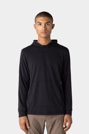 A young man with short hair wears the 686 Men's Everywhere drirelease® Long Sleeve Hooded Tech Tee in black and light brown pants, standing against a plain light gray background, facing forward with a neutral expression.