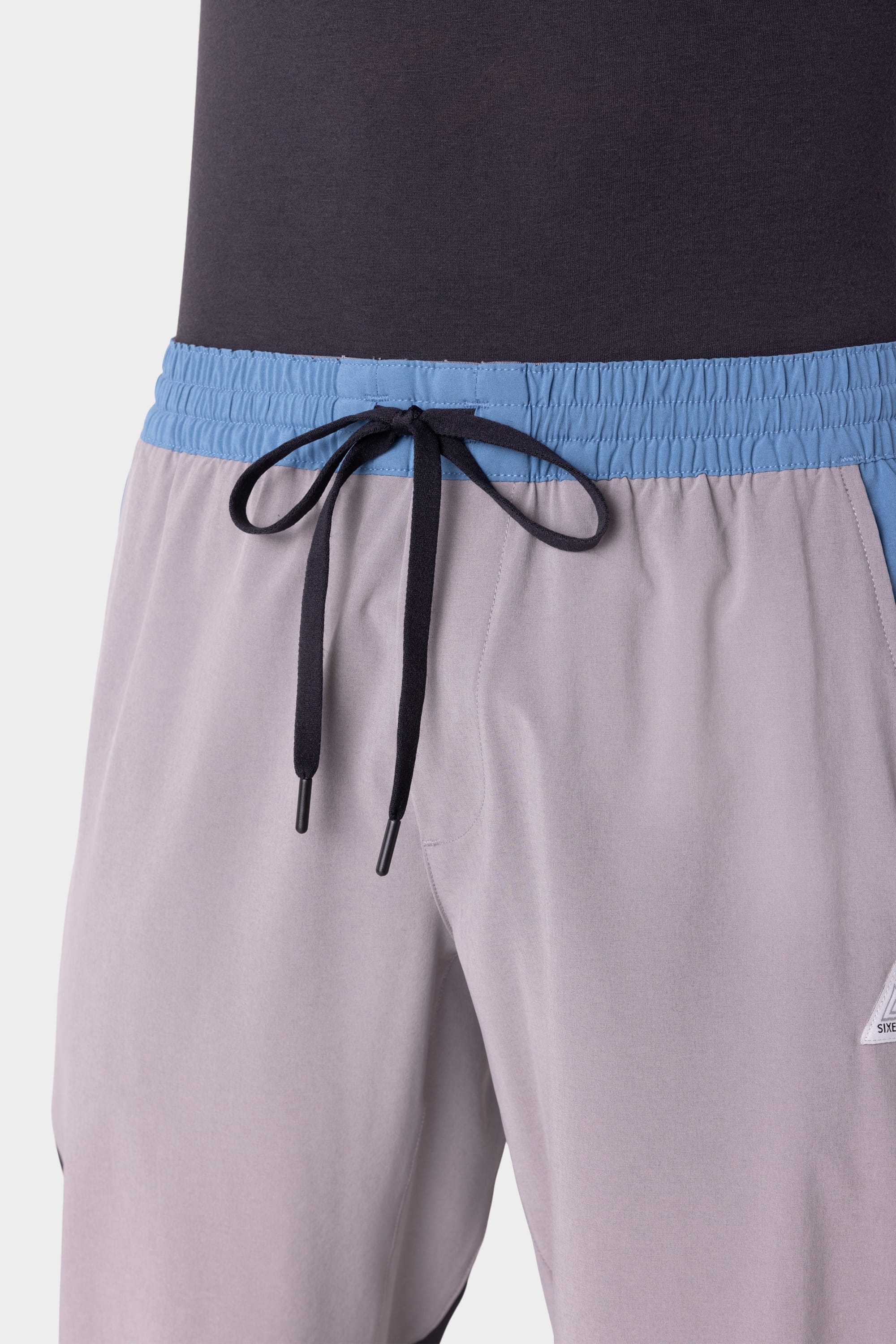 A close-up of a person wearing the 686 Men's ATP Stretch Performance Pant in light gray with a blue waistband and black drawstring, paired with a dark gray shirt. Part of the 686 logo is visible on the pant.
