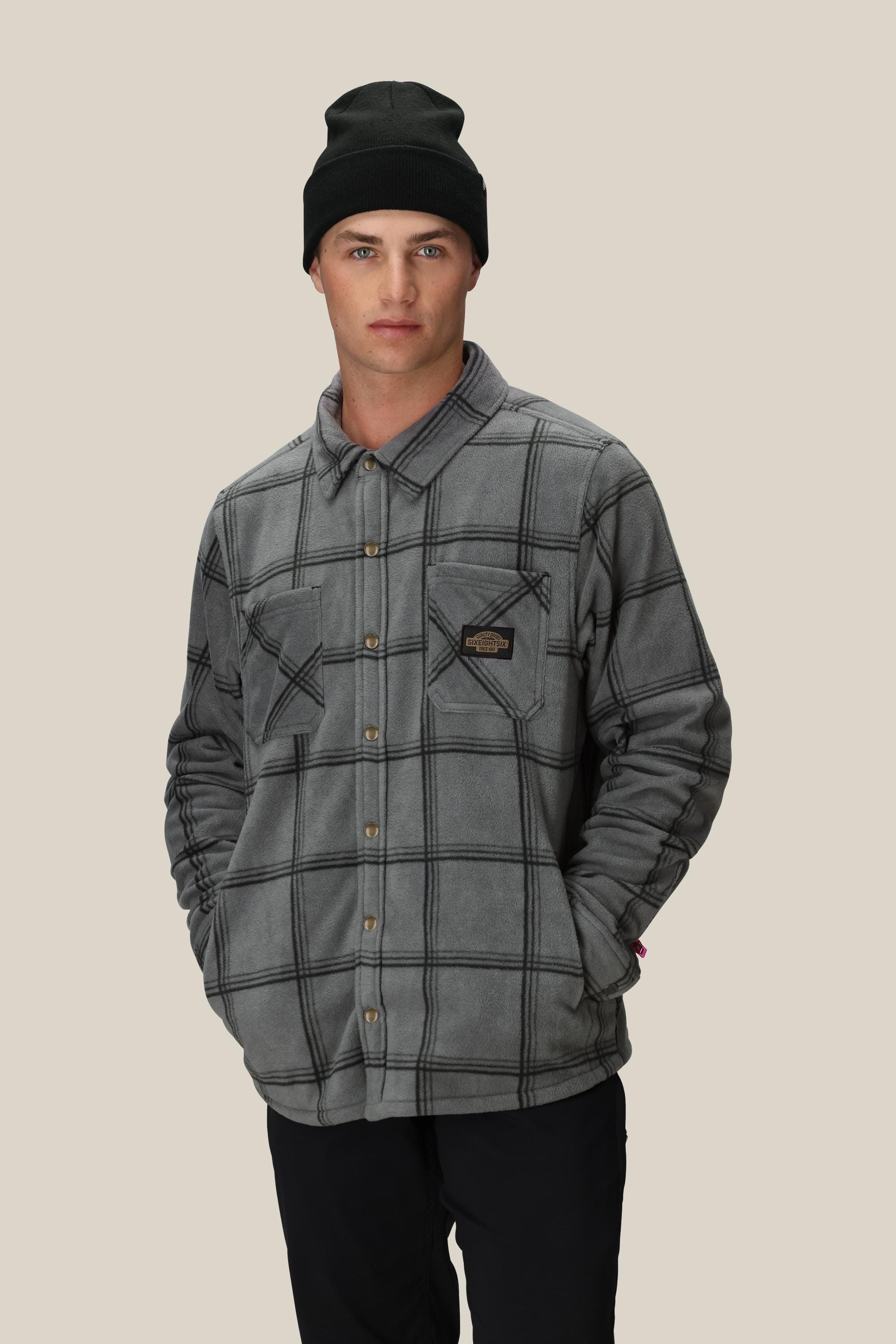 A man stands against a light background with his hands in his pockets, wearing a black beanie, black pants, and the 686 Mens Sierra Fleece Flannel by 686.