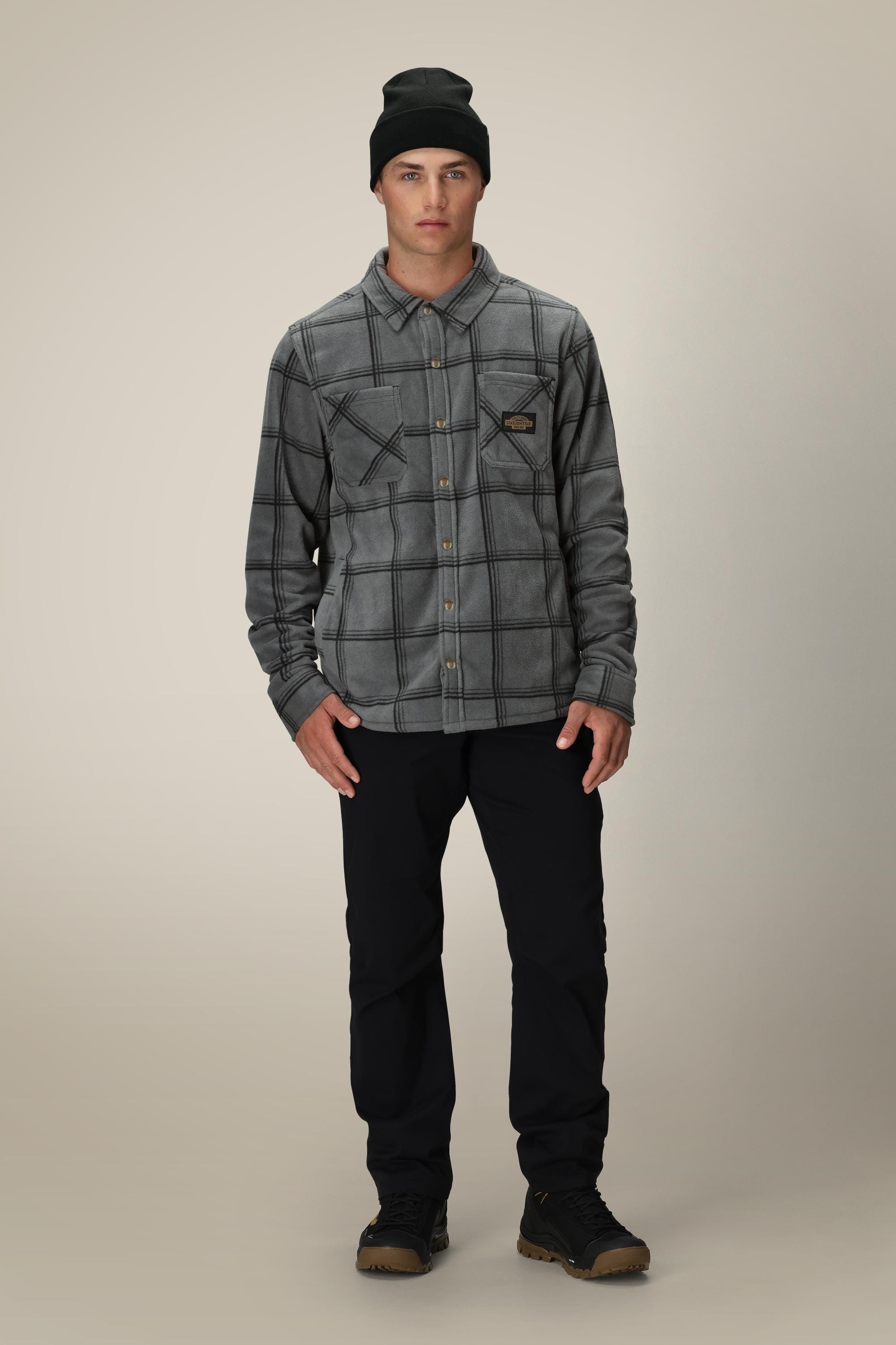 A young man faces forward against a beige background, layering the 686 Mens Sierra Fleece Flannel over black pants and shoes.