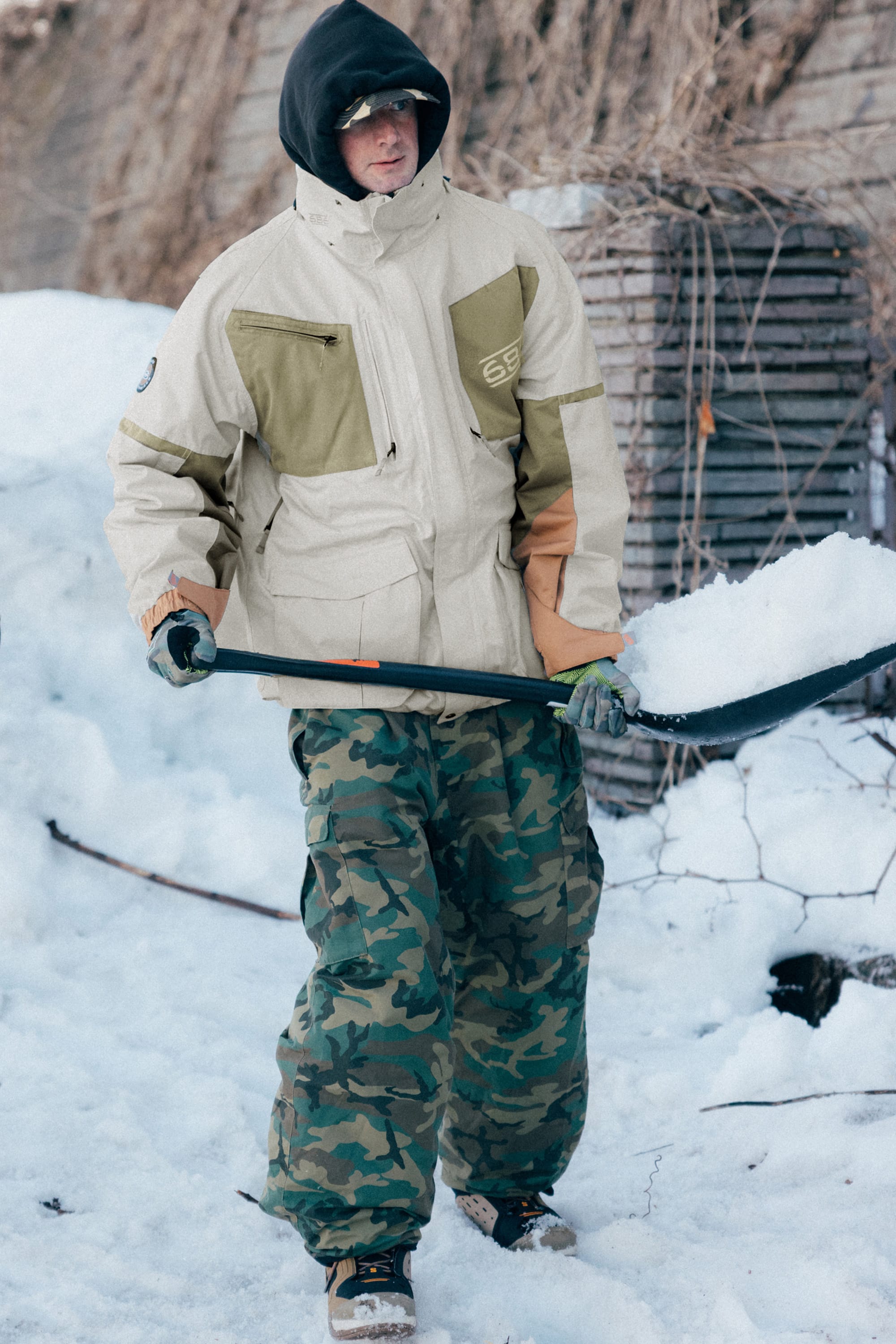 A person wearing the 686 Mens 2001 Transfer Jacket stands outdoors in the snow with a shovel. Camouflage pants, bare branches, and stacked pallets are visible in the background.