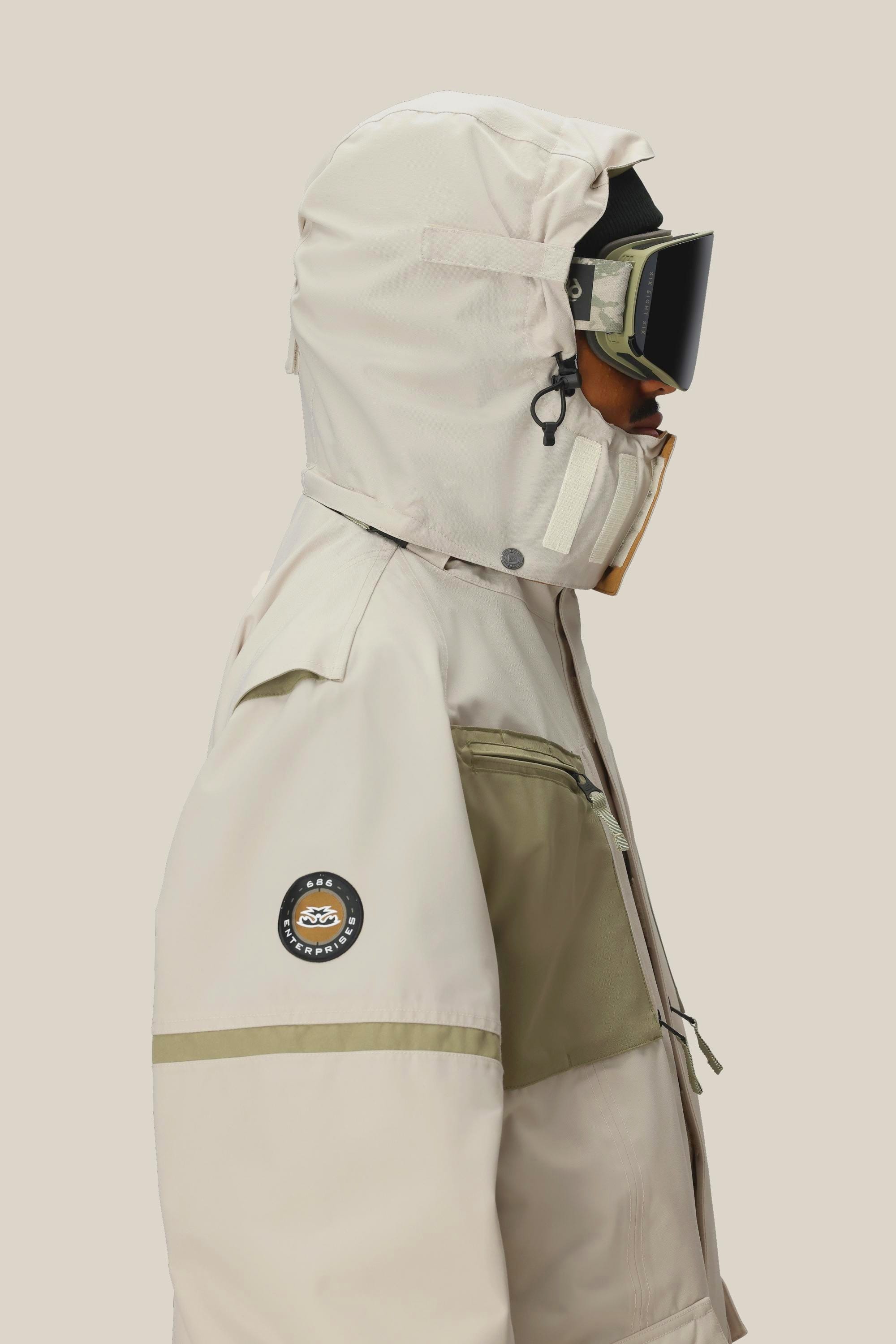 A person is shown in profile against a plain background, wearing the 686 Mens 2001 Transfer Jacket with a sleeve patch, plus ski goggles and a face covering.