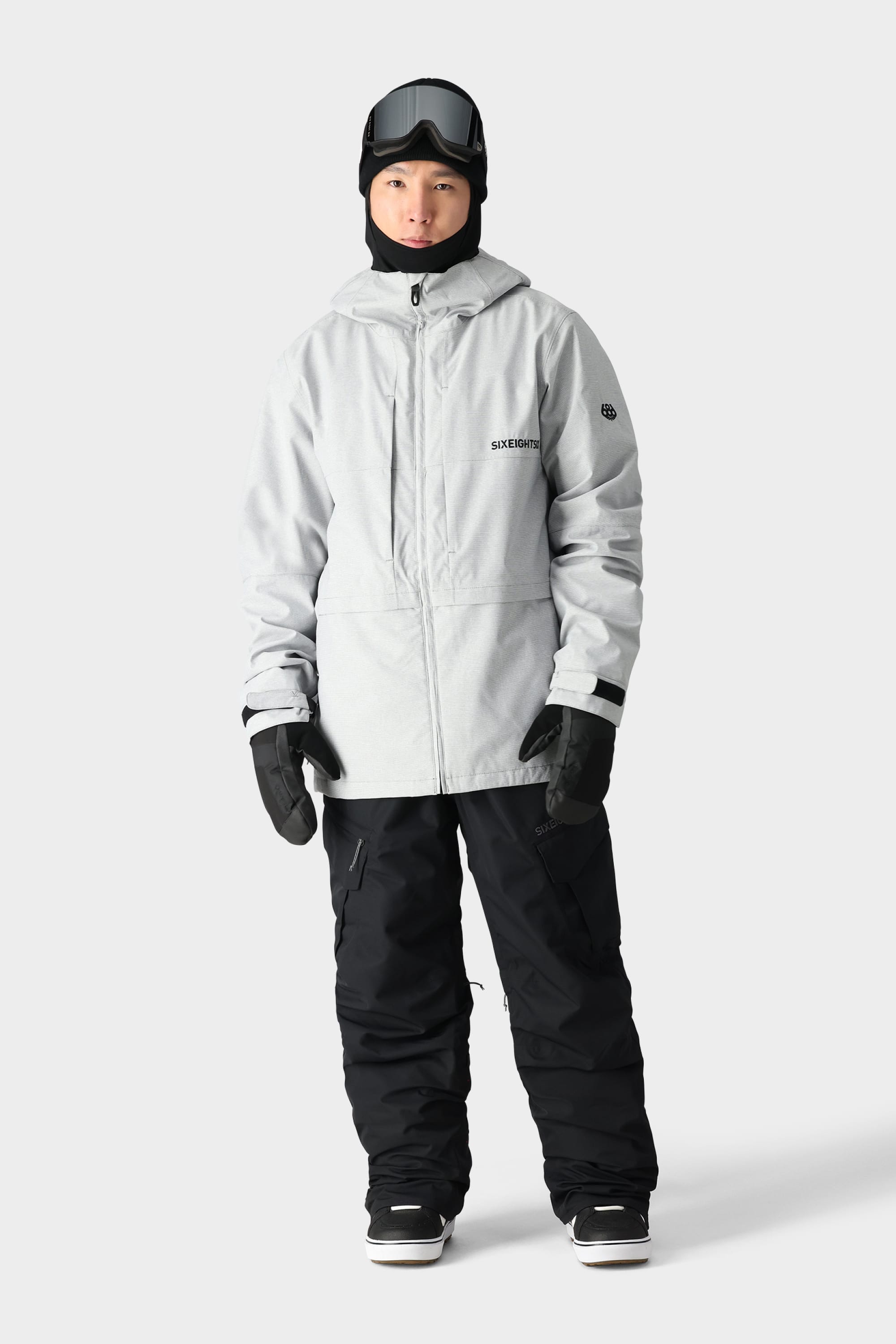 A person wearing a light gray hooded winter jacket, 686 Men's SMARTY® 3-in-1 Cargo Bib from 686, black gloves, snow boots, a black balaclava, and ski goggles stands against a plain light gray background.