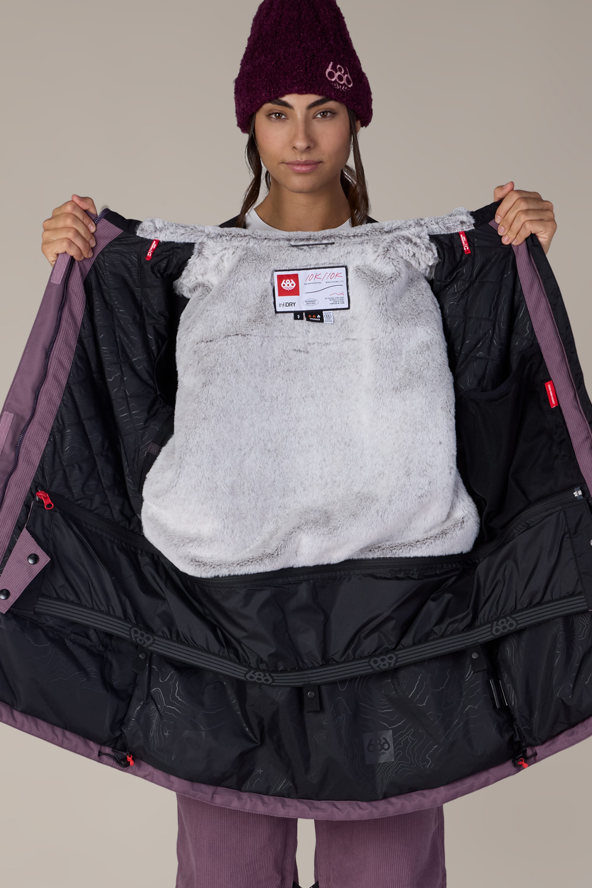 A woman wearing the 686 Women’s Spirit Insulated Jacket in purple and black opens it to reveal its soft, gray inner lining and interior label, standing against a plain, light background.