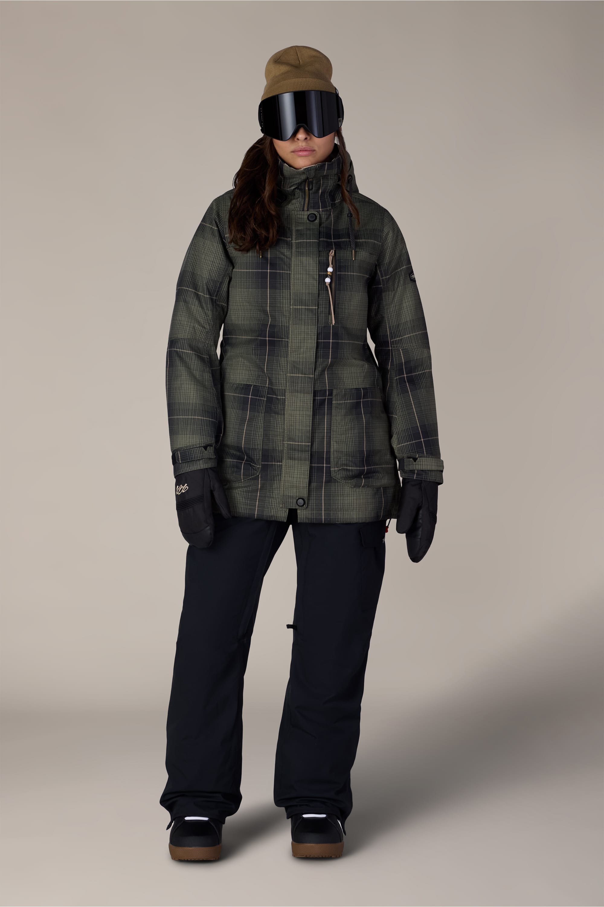 A person is shown facing forward against a plain background wearing the 686 Women’s Spirit Insulated Jacket by 686, along with black snow pants, gloves, boots, ski goggles, and a tan hat.