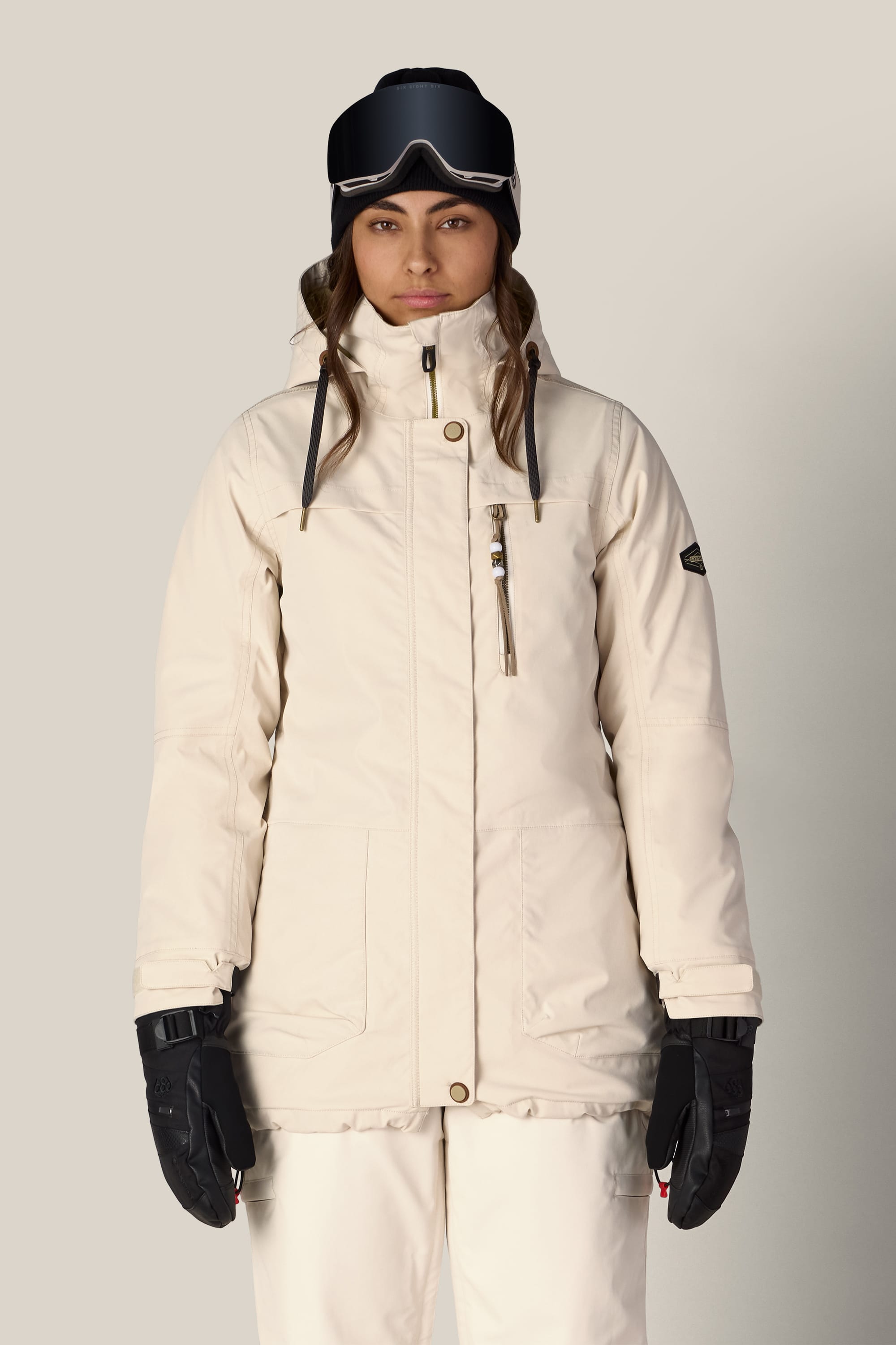 A woman wearing the 686 Women’s Spirit Insulated Jacket by 686 stands facing forward against a plain light background, paired with white pants, black gloves, and ski goggles.