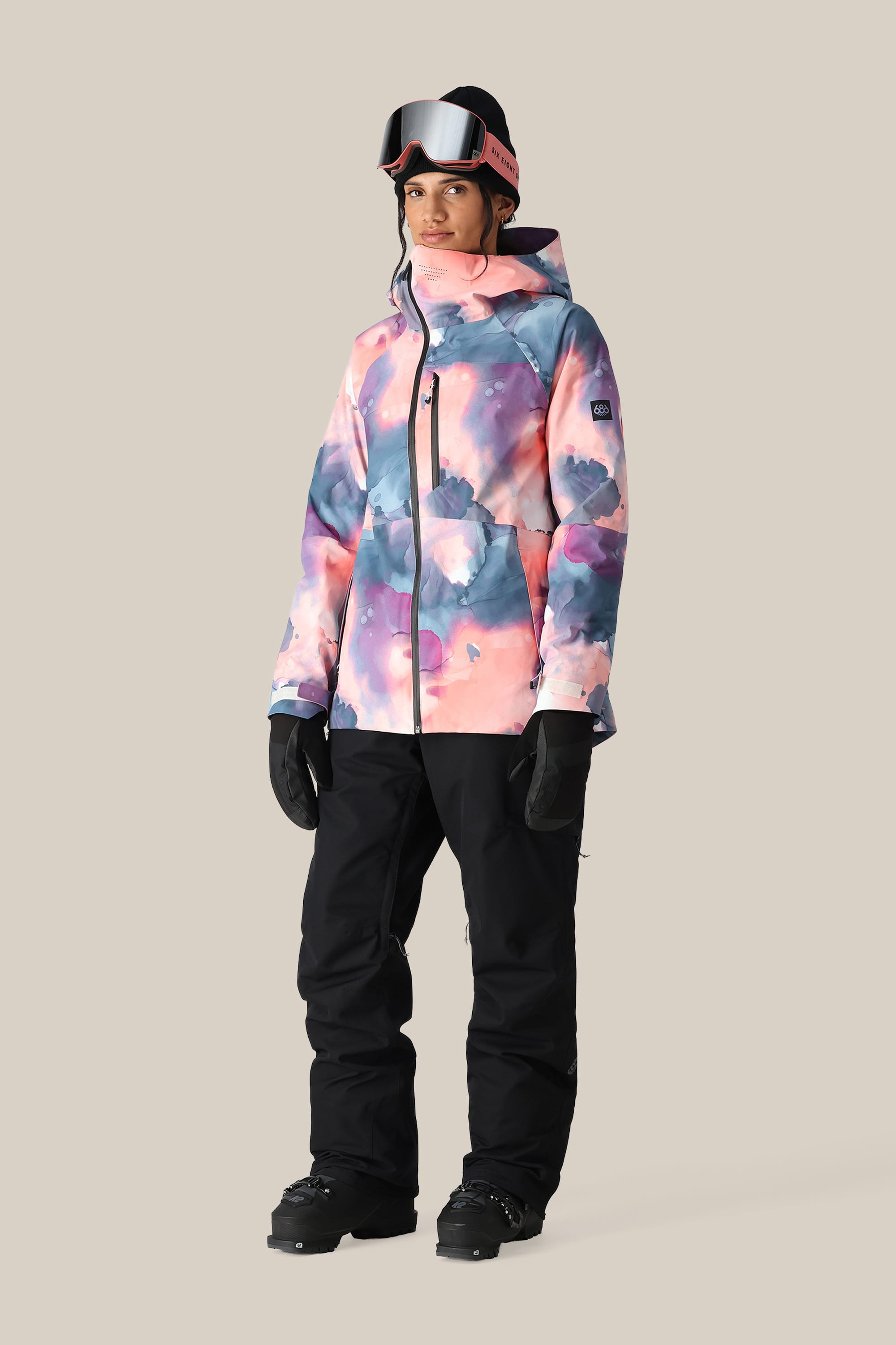 A person wearing a colorful pastel tie-dye ski jacket, 686 Women's SMARTY® 3-in-1 Cargo Pant by 686 with a removable sweatpant liner, black gloves, and ski goggles stands against a plain beige background.