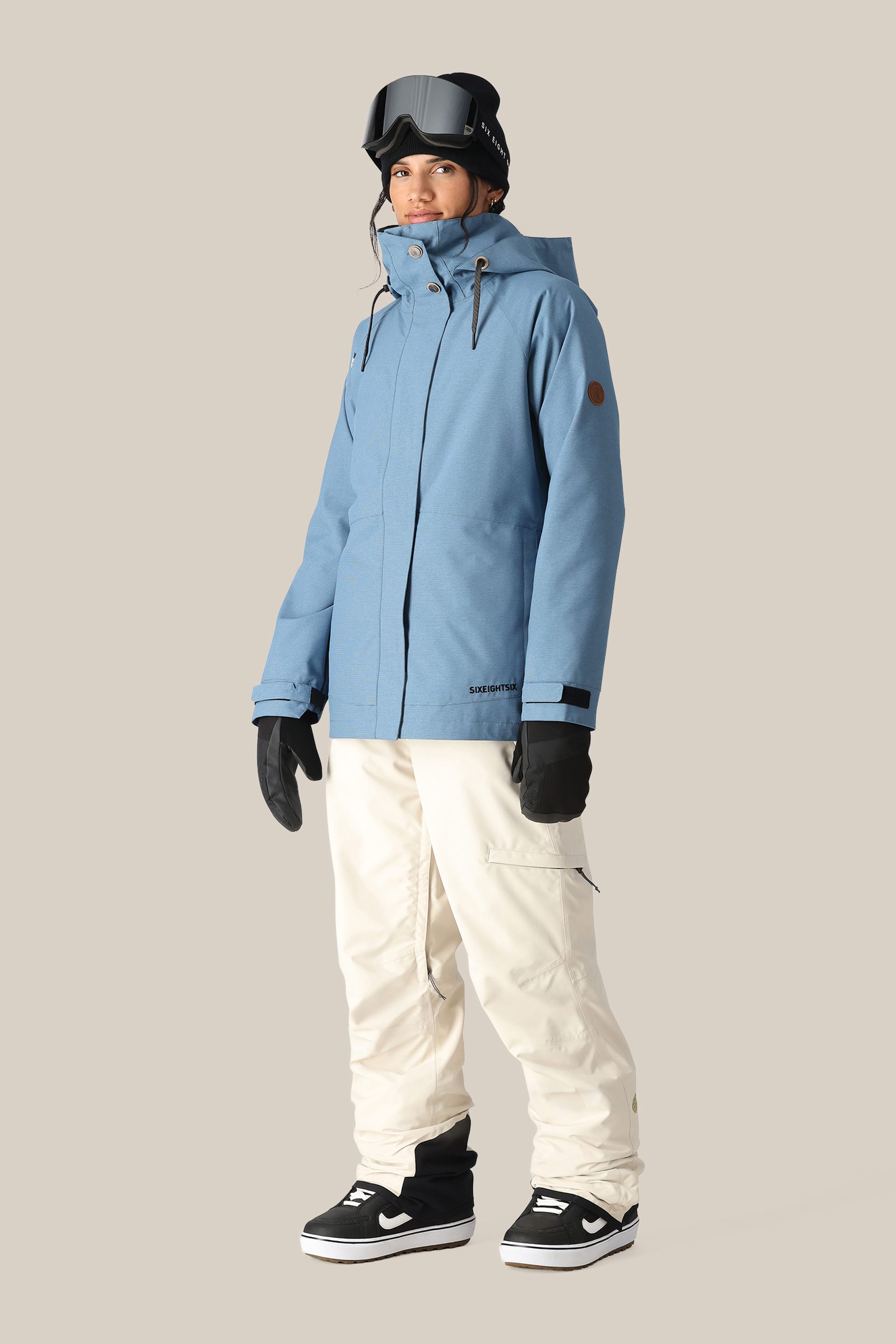 A person wearing white 686 Women's SMARTY® 3-in-1 Cargo Pant by 686, a blue hooded winter jacket, black gloves, black snow goggles, and black sneakers stands against a plain beige background.