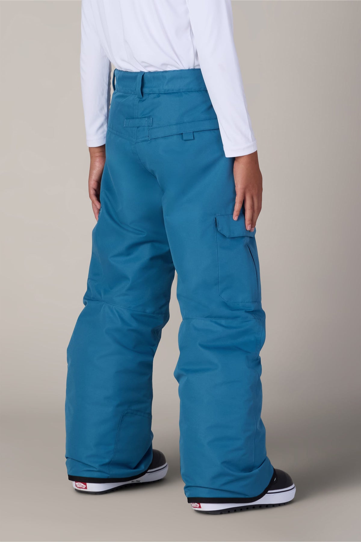 A person is shown from the back against a plain background, wearing the 686 Boys Infinity™ Cargo Insulated Pant in a loose-fitting blue style, paired with a white long-sleeve shirt and black-and-white snow boots.