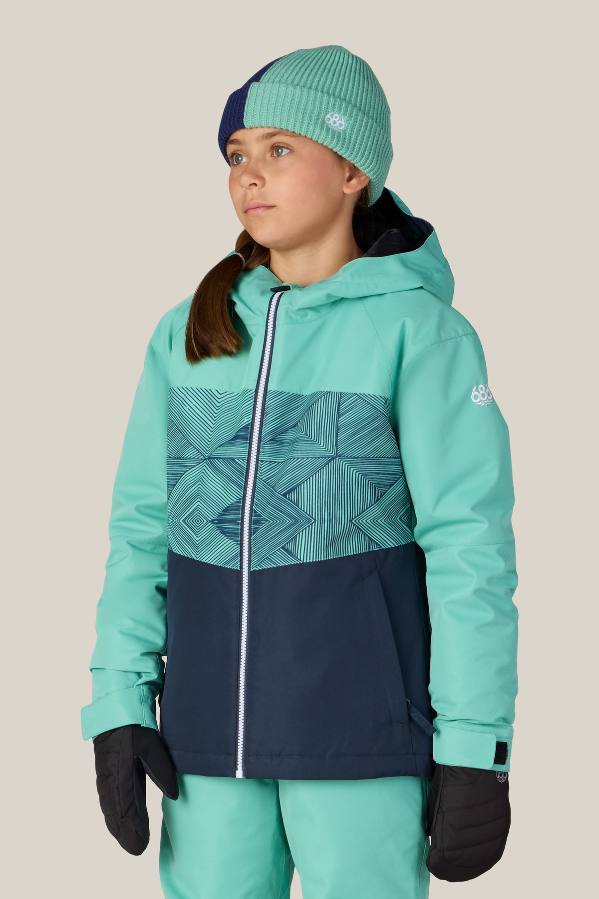 A girl wearing the 686 Girls’ Athena™ Insulated Jacket in teal and navy, paired with matching teal pants, black gloves, and a green beanie with a navy section, stands against a plain light background, looking slightly to the side.