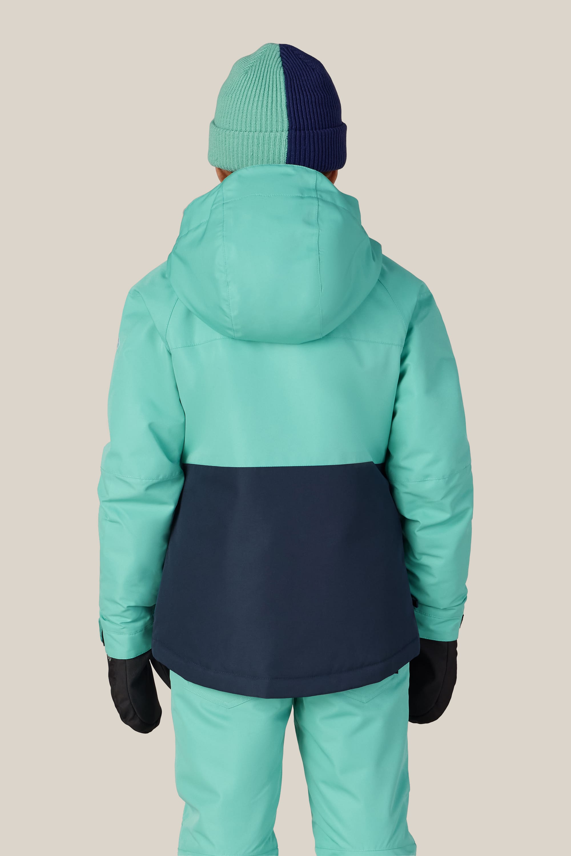 A child stands facing away against a plain beige background, wearing the 686 Girls’ Athena™ Insulated Jacket in teal and navy by 686, along with matching teal pants, black gloves, and a two-toned teal and navy knit beanie hat.
