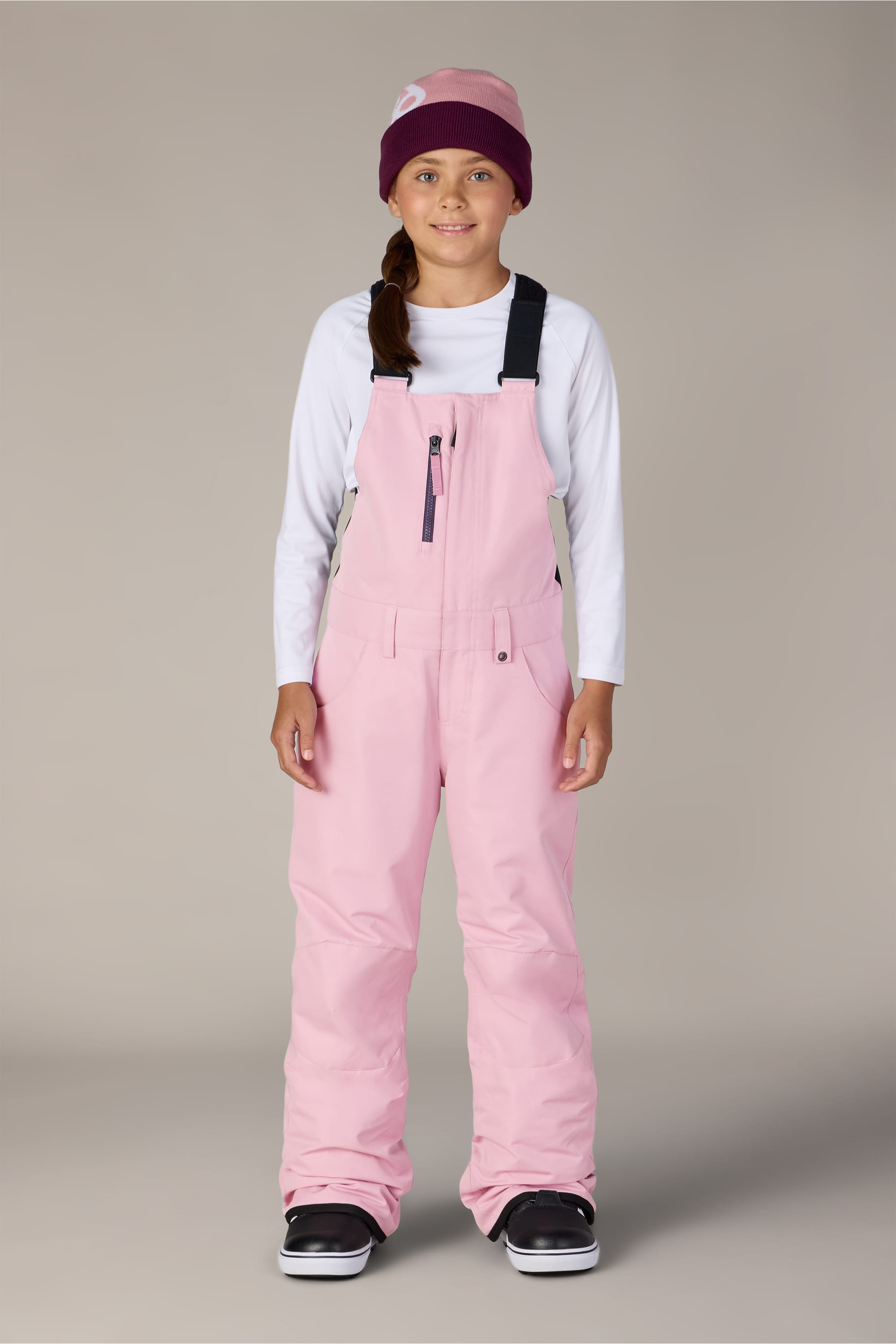 A young girl stands facing forward against a plain light gray background, wearing a white long-sleeve shirt and the 686 Girls Sierra Insulated Bib snow pants in light pink—crafted from waterproof polyester bib fabric by 686—paired with a maroon beanie and black snow boots.