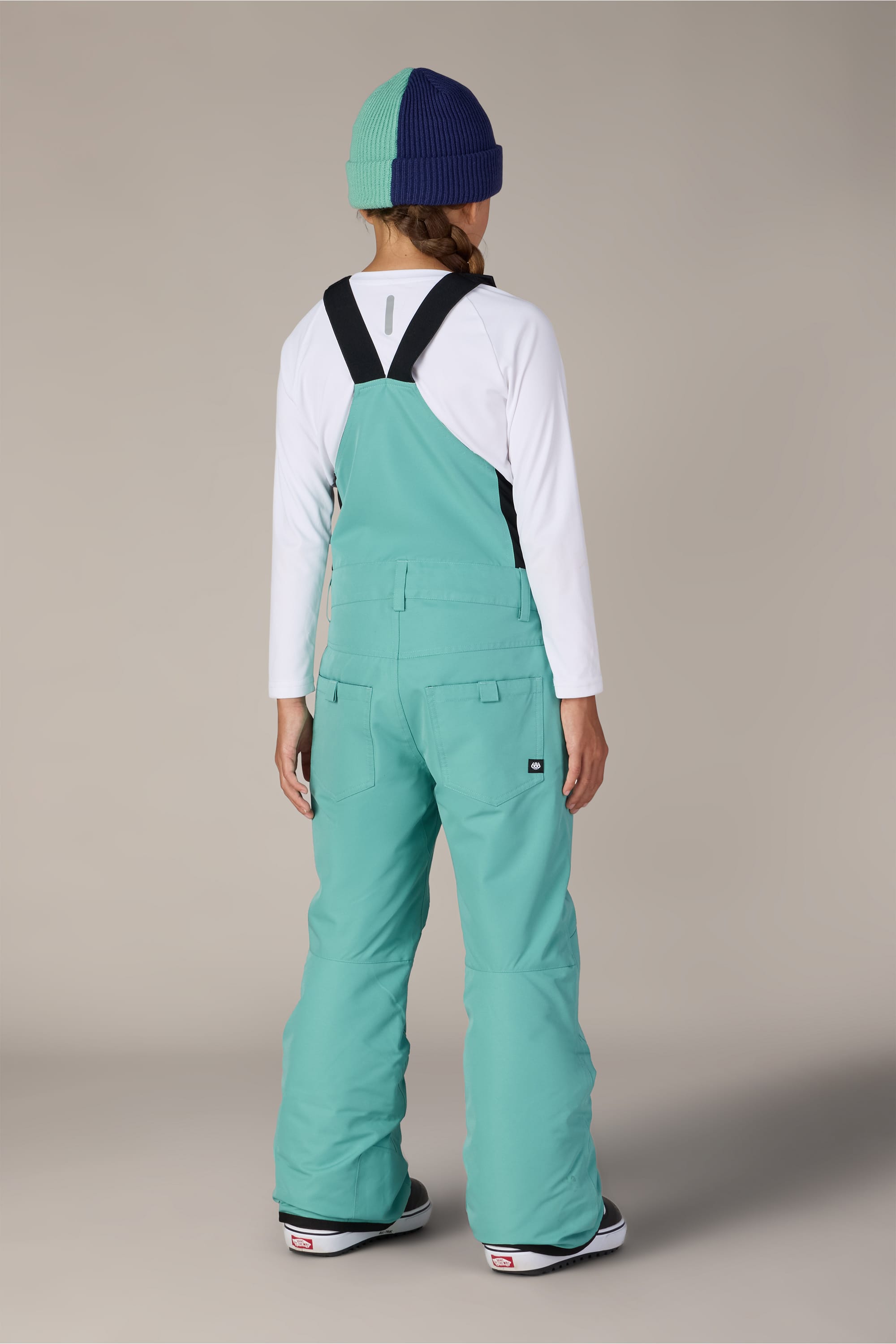 Facing away, the person is wearing the 686 Girls Sierra Insulated Bib from 686 in an aqua hue, styled in a polyester waterproof bib design. The look is completed with a long-sleeve white shirt, two-toned knit beanie, and snow boots, all set against a simple neutral backdrop.