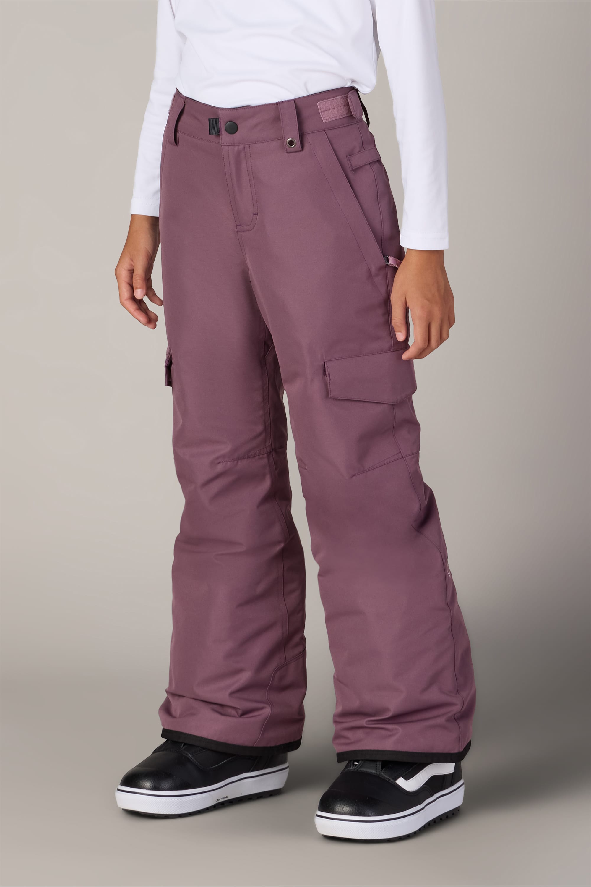 A person is shown from the waist down wearing 686 Girls’ Lola Insulated Pant in mauve, which features light insulation and cargo pockets, paired with a white long-sleeve shirt and black snowboard boots. The individual stands against a plain light gray background.