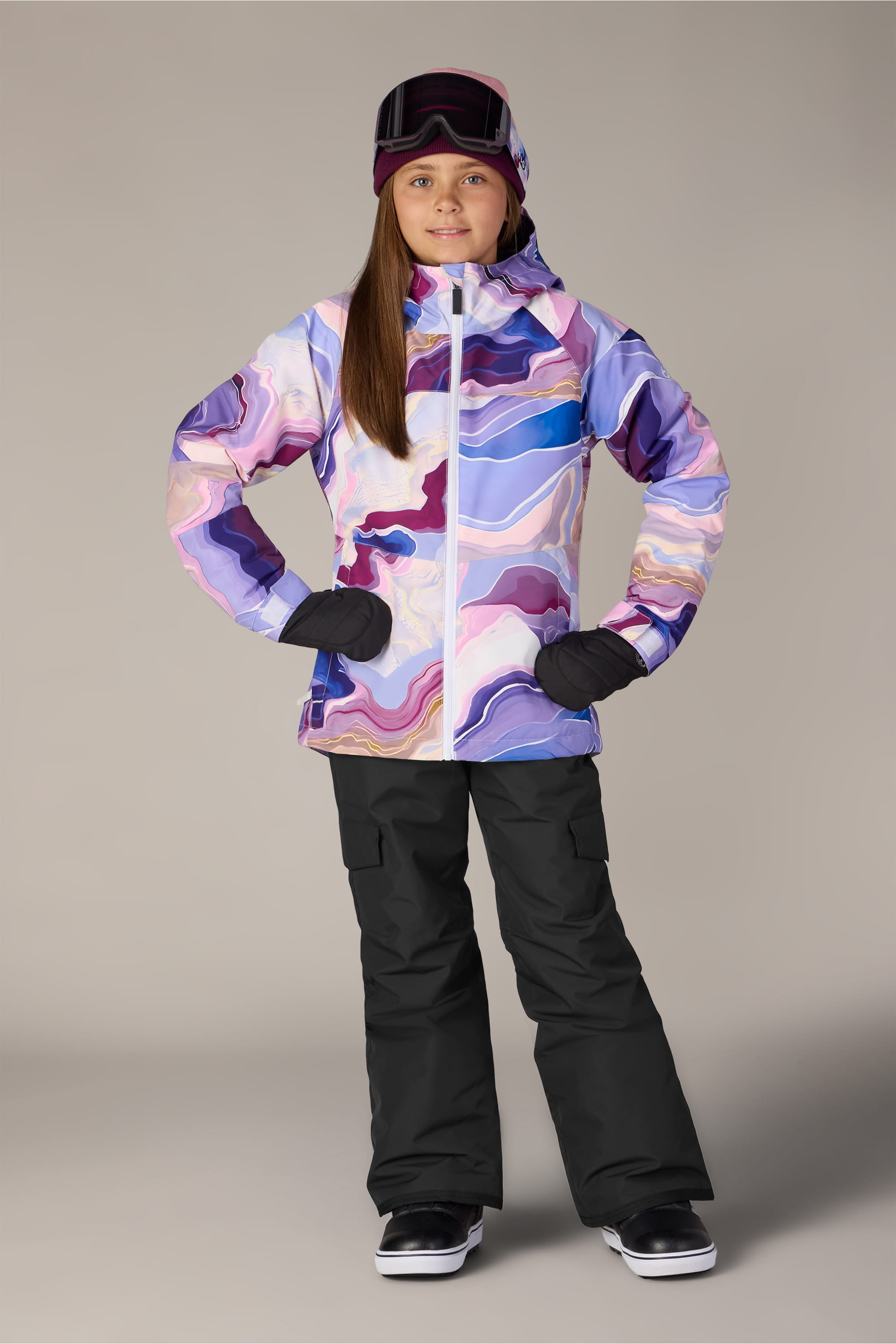 A person wearing a colorful, abstract-patterned winter jacket with light insulation, 686 Girls’ Lola Insulated Pant in black, black gloves, and a ski helmet with goggles stands with hands on hips against a plain background.