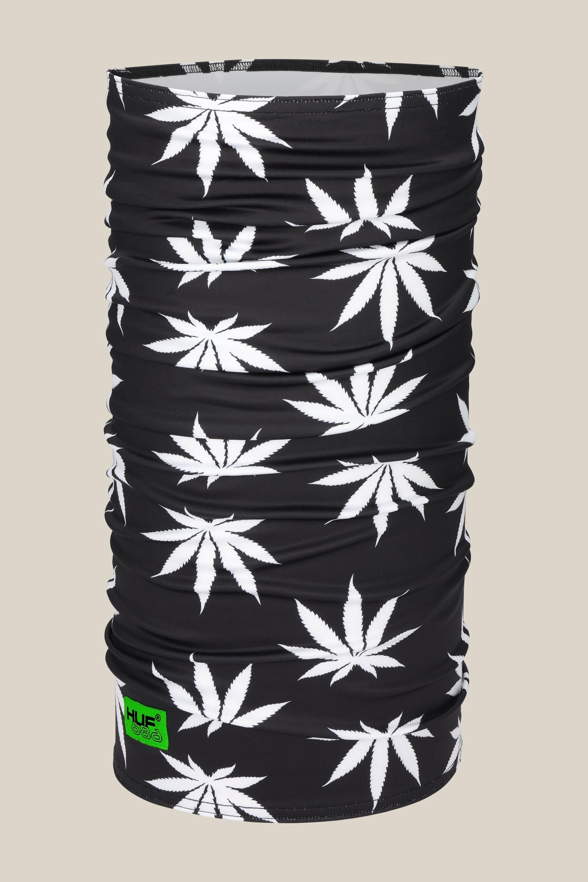 The 686 Double Layer Face Warmer by 686 is a black neck gaiter with a repeating white cannabis leaf pattern and a small neon green logo tag. The solid beige background pairs well with both high-waist and low-waist outfits.