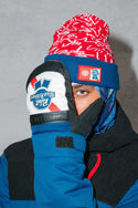 A person in a red knit beanie and blue jacket holds up a white 686 Mens infiLOFT Recon Mitt by 686, partially covering their face, while looking at the camera against a plain gray background.