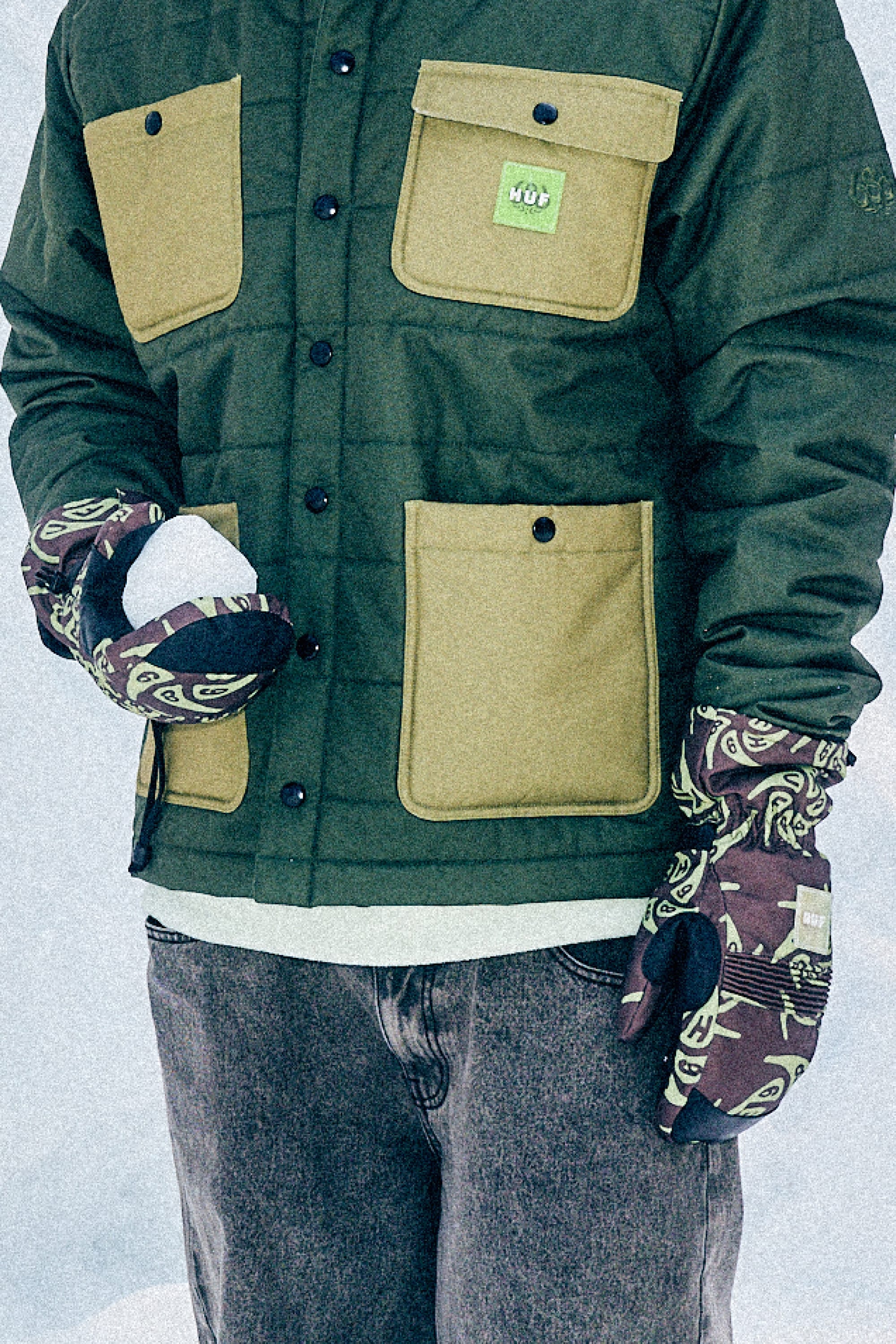 A person wearing a green and tan jacket, jeans, and 686 Lander™ Mitt gloves by 686 holds a snowball in one hand while standing outdoors in the snow.