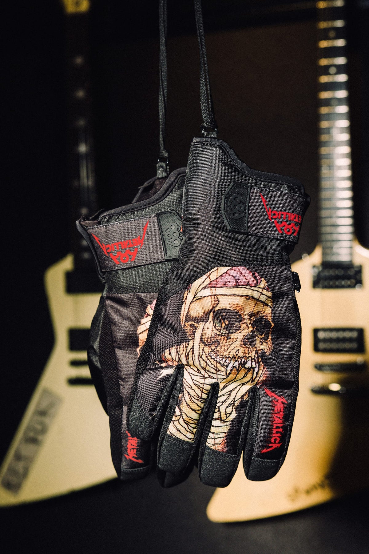 686 Men’s Primer Gloves in black featuring a mummy-wrapped skull graphic and Metallica logos, hanging in front of electric guitars.