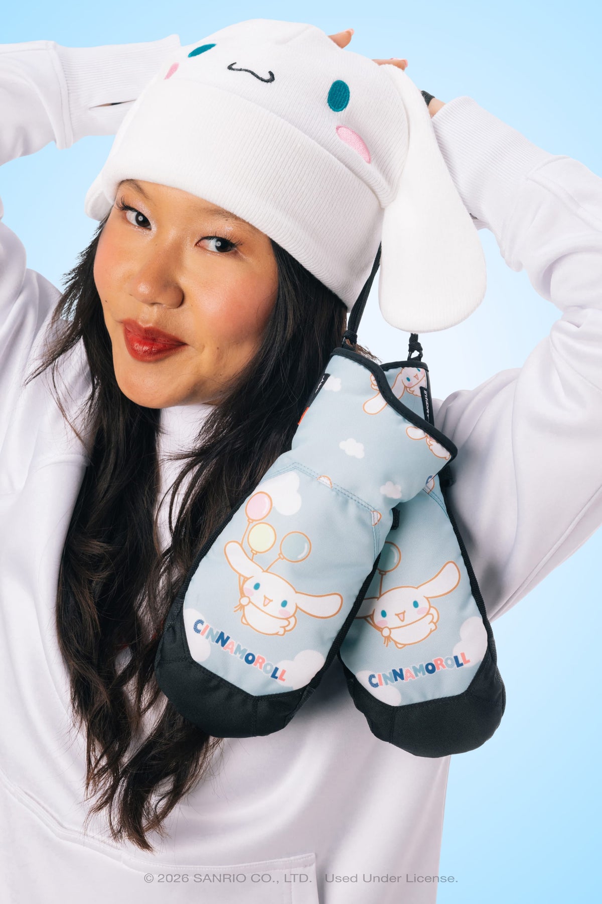 A person wearing a white bunny hat and holding 686 Women’s Revel Mitts by 686, smiling in front of a blue background.