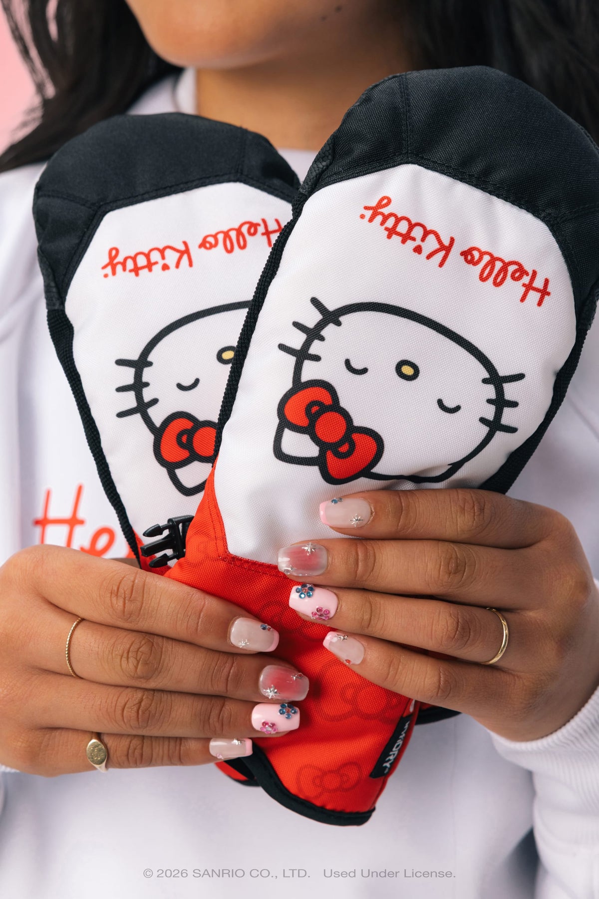 A person holding a pair of 686 Women’s Revel Mitts by 686, featuring a Hello Kitty theme with red bows. The person has light pink nails adorned with floral designs, is wearing gold rings, and is dressed in a white sweatshirt.