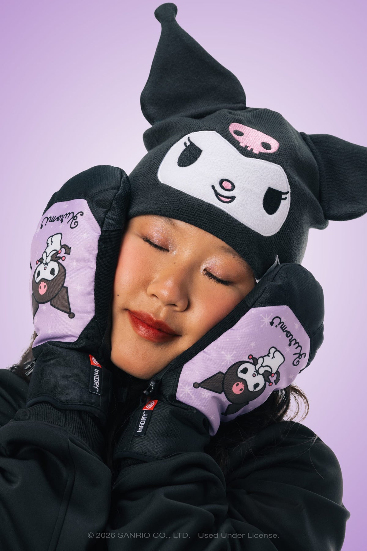 A woman wearing a black Kuromi hat and matching purple 686 Women’s Revel Mitts by 686 smiles softly with her eyes closed, resting her face in her hands against a light purple background.