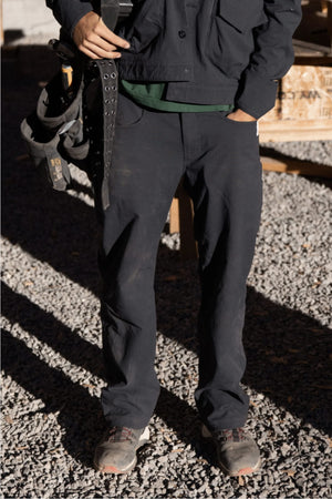 A person stands outdoors on gravel wearing the 686 Men's Unwork® Pant - Relaxed Loose Fit, a work jacket, and sturdy shoes. With a tool belt at their waist and one hand in their pocket, their face is not visible.