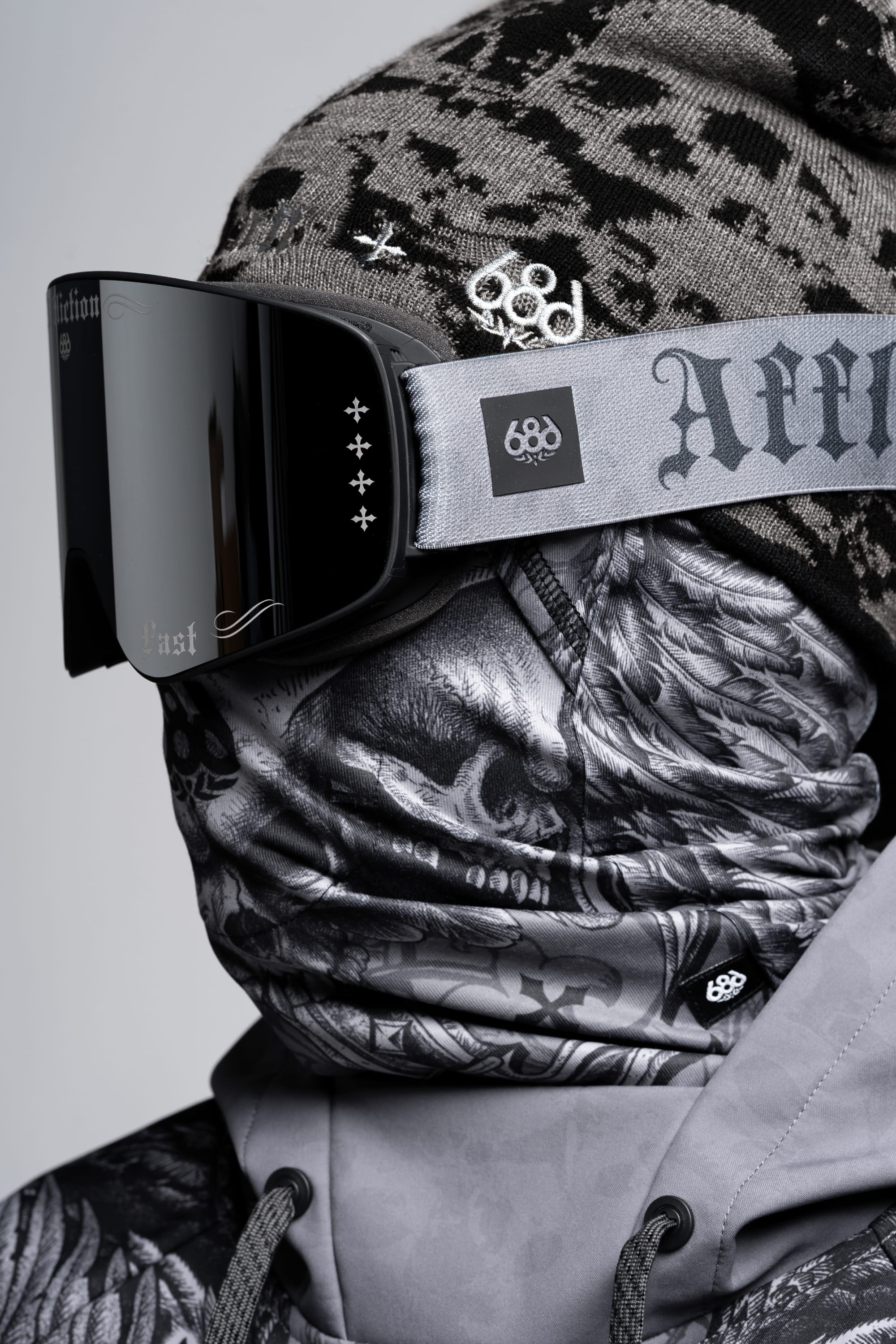 A person wears a patterned grey beanie, a 686 SMARTY® Magnetic Goggle by 686, and a black and white skull scarf covering their face and neck. Their outfit also includes a grey hoodie with visible brand logos.