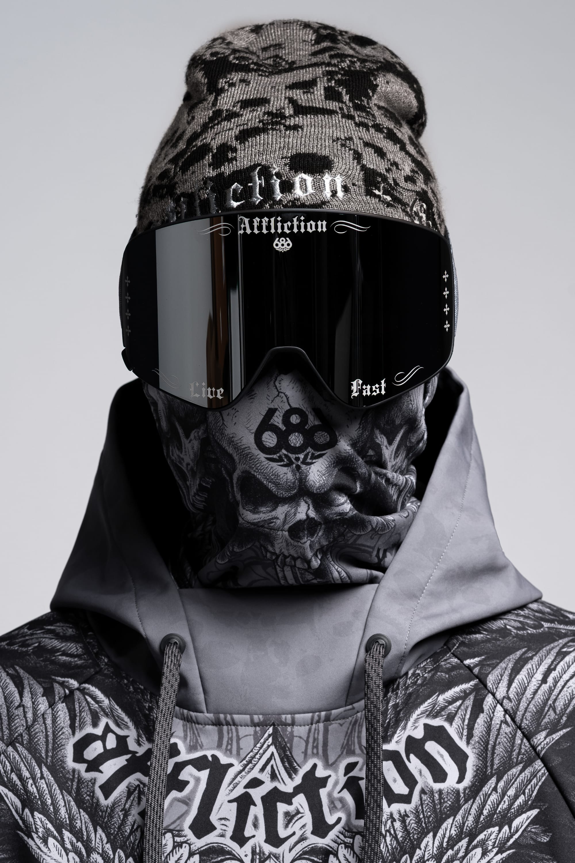 A person wearing a patterned beanie, 686 SMARTY® Magnetic Goggle, and a skull-printed face covering—all branded with Affliction. The hoodie displays bold, dark designs and “Affliction” across the chest.