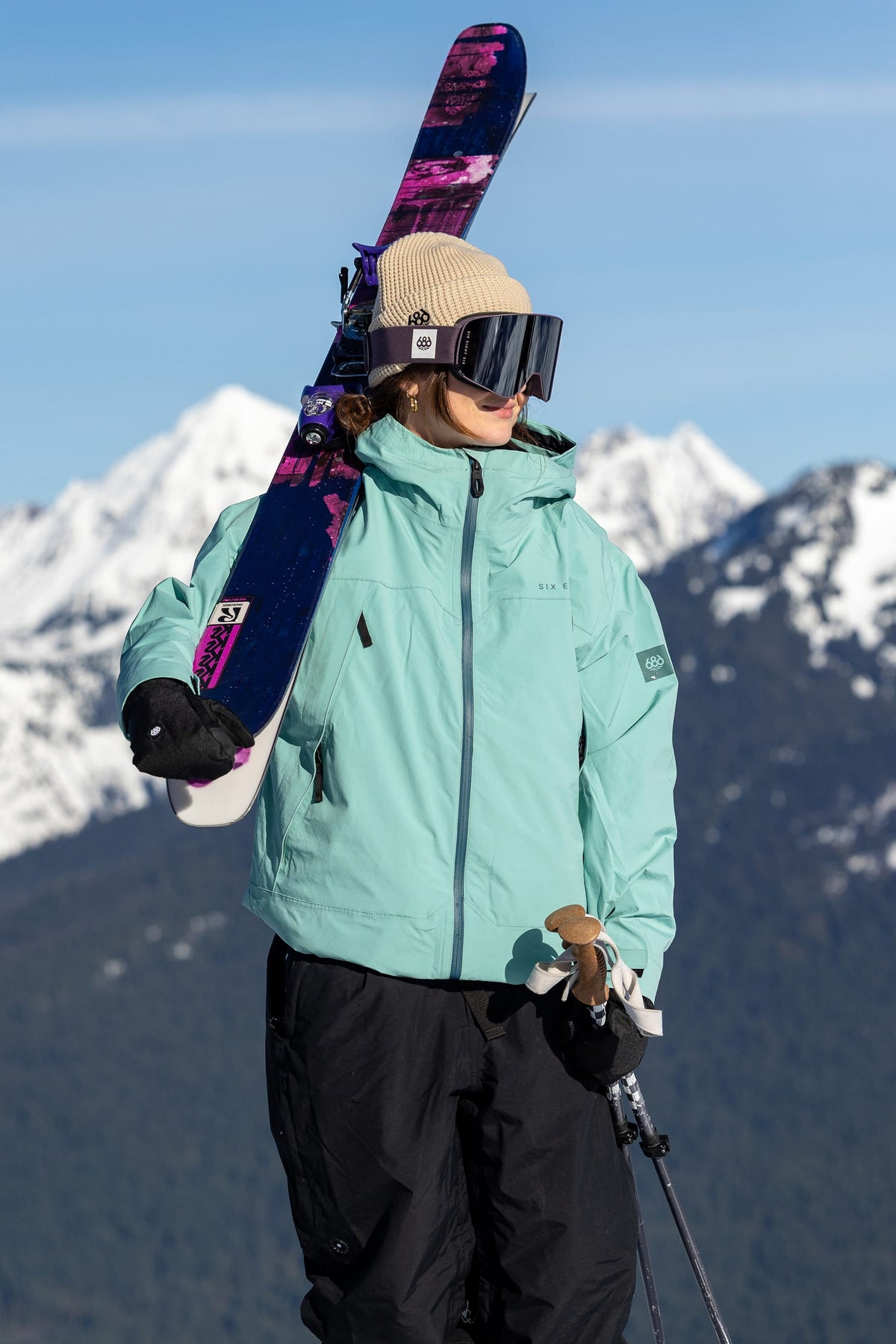 Wearing the 686 SMARTY® Magnetic Goggle from 686, a skier in a light blue jacket, black pants, and beige beanie stands with skis over their shoulder in front of snowy mountains on a sunny day.
