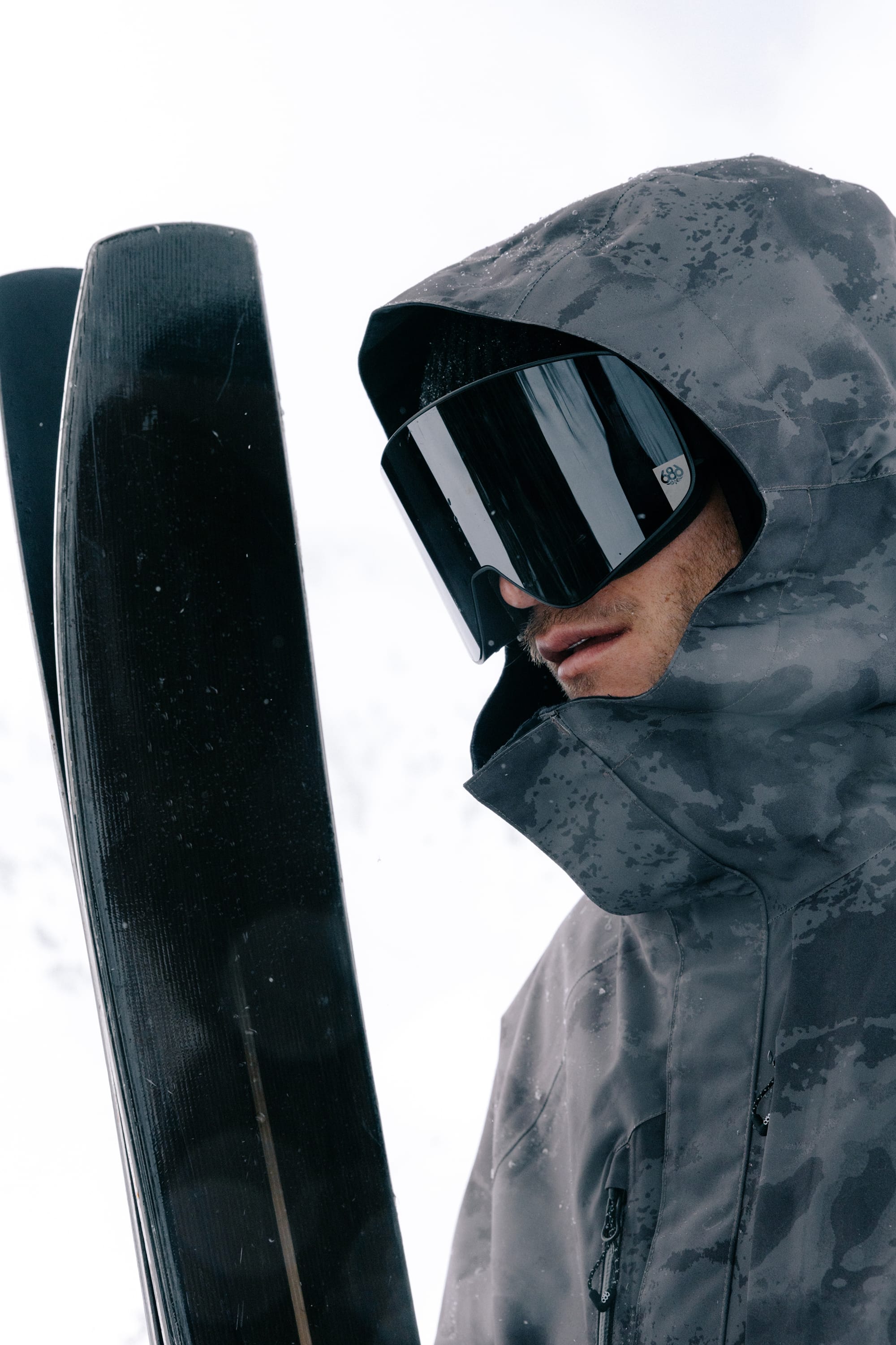 A person wearing a gray patterned hooded jacket and the 686 SMARTY® Magnetic Goggle by 686 holds a pair of black skis upright against a snowy, overcast background.