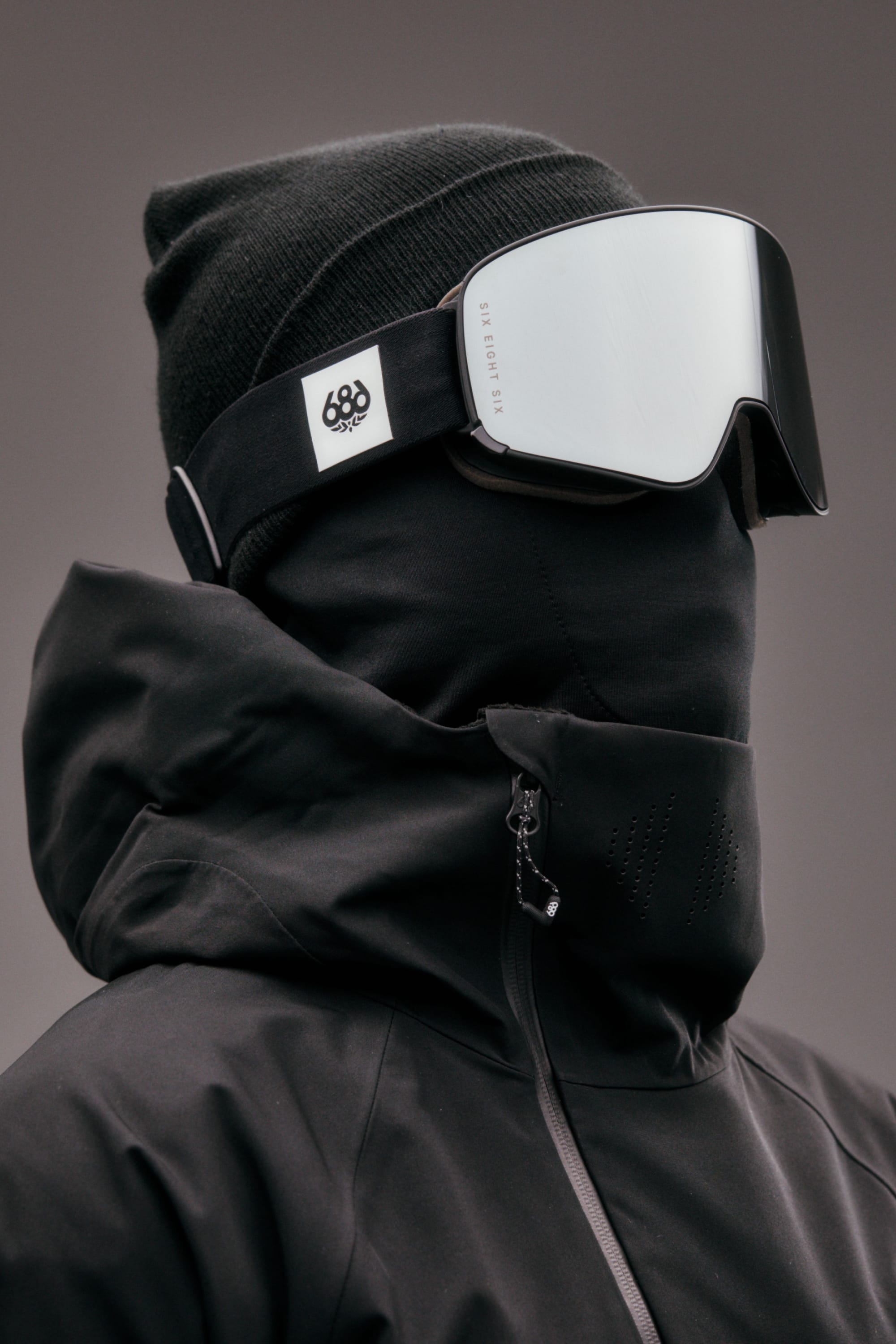A person wearing a black beanie, the 686 SMARTY® Magnetic Goggle by 686 with interchangeable lenses, and a high-collar black jacket that fully covers their face and neck, stands against a plain gray background.