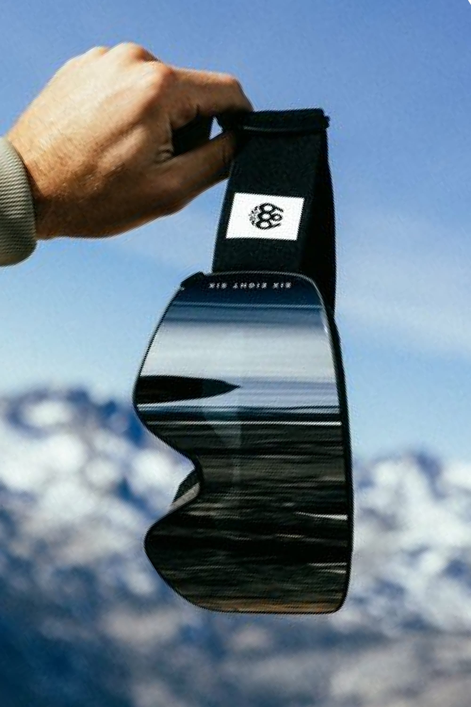 A hand holds up the 686 SMARTY® Magnetic Goggle, which reflects a blurred landscape, with snow-covered mountains and a blue sky visible in the background.