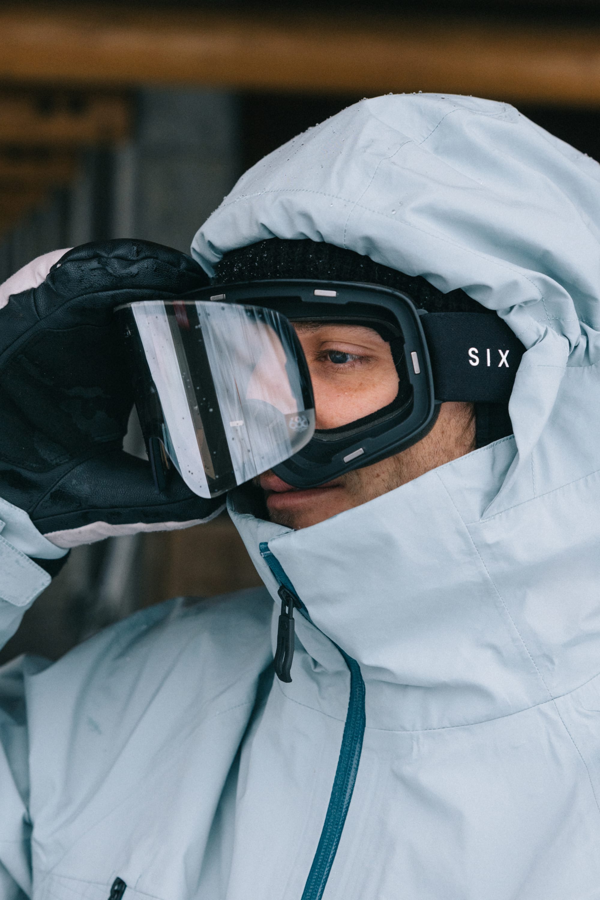 A person in a light blue winter jacket and black 686 SMARTY® Magnetic Goggle by 686 looks into the distance, adjusting the goggles with a gloved hand.