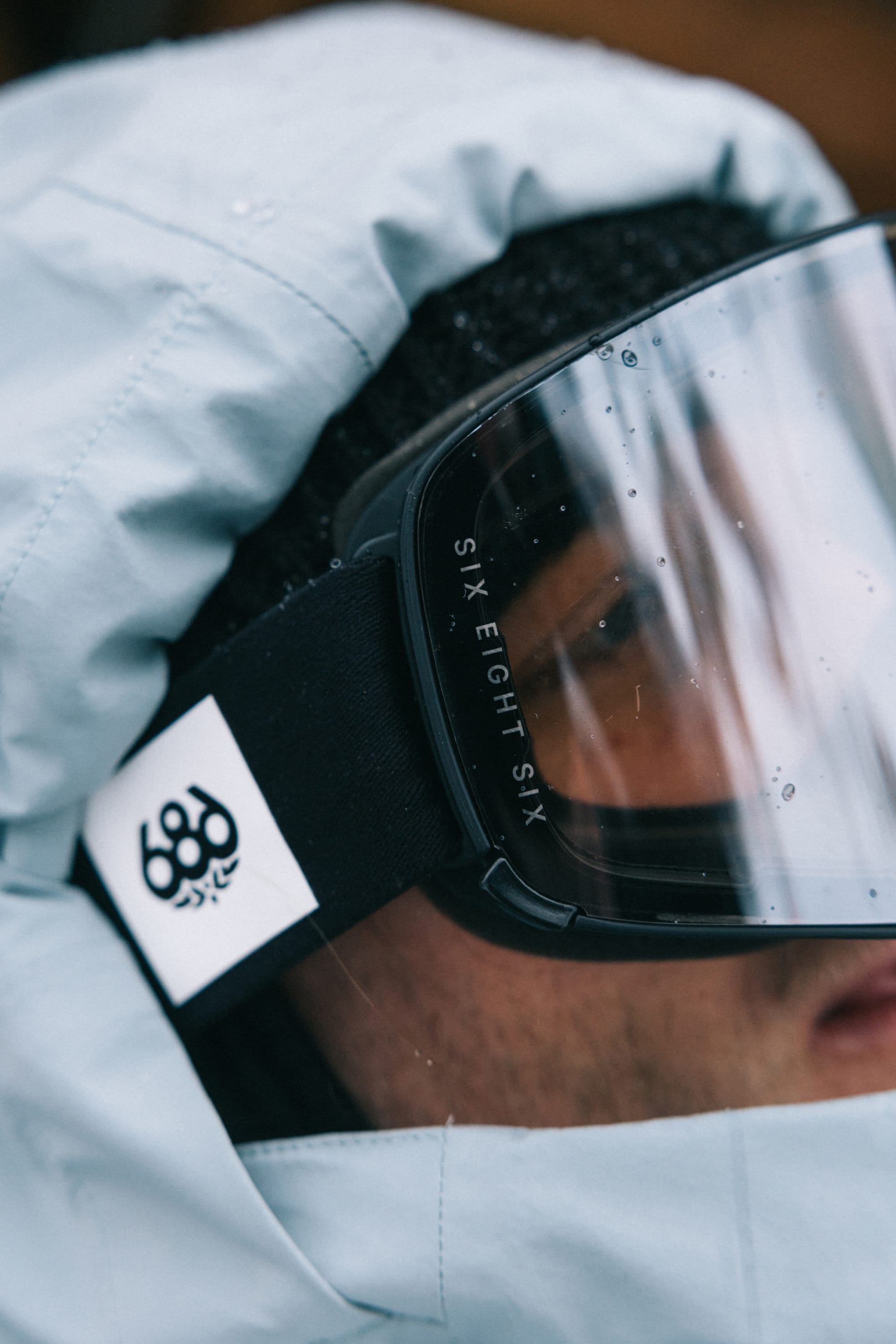 A close-up of a person wearing a light blue hooded jacket, black beanie, and the 686 SMARTY® Magnetic Goggle with “686” branding visible on the strap. The focus is on the goggles and their branded strap.