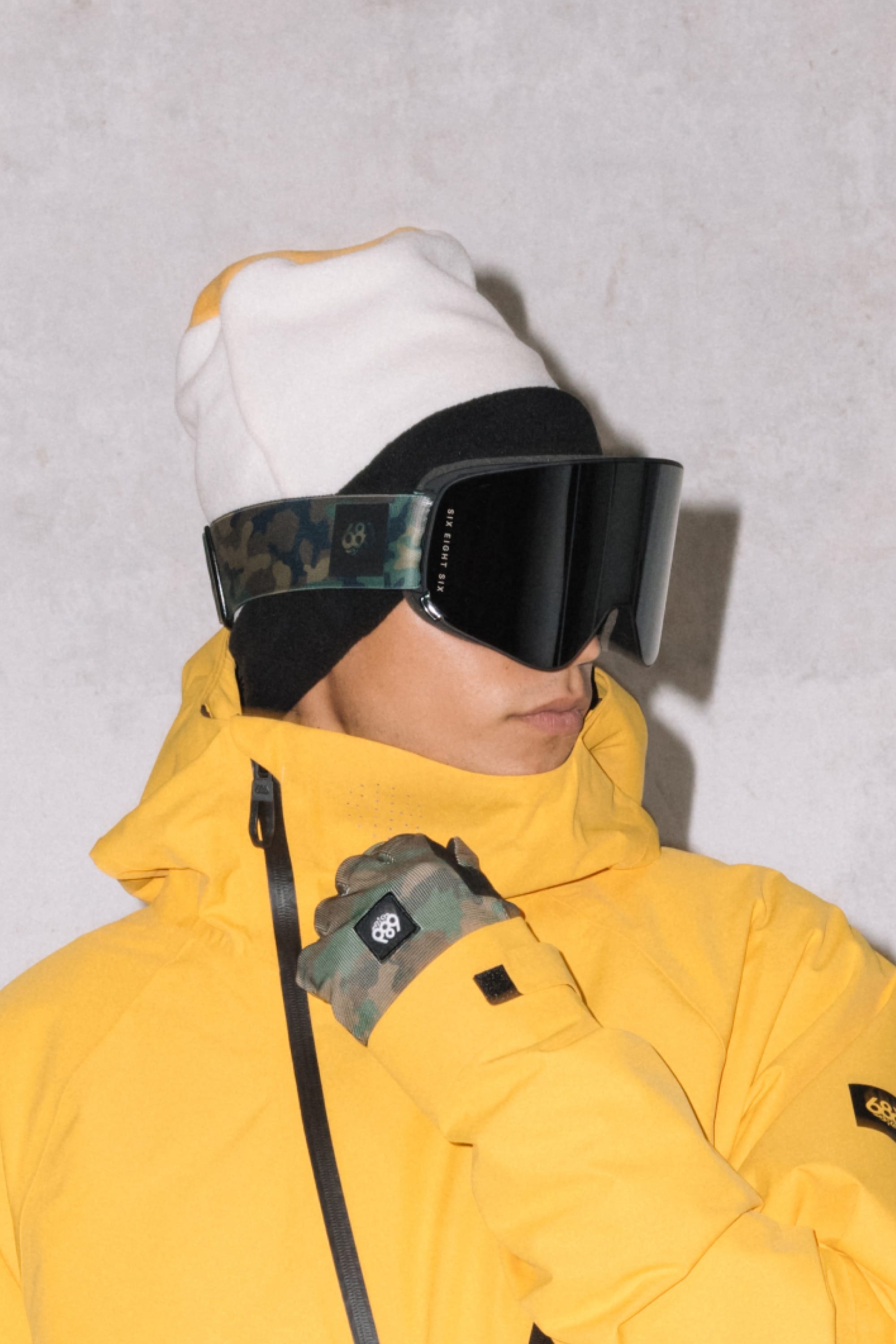 A person in a yellow winter jacket, black and white beanie, and 686 SMARTY® Magnetic Goggle with a camouflage strap stands against a light gray background, finishing the look with matching camouflage gloves.