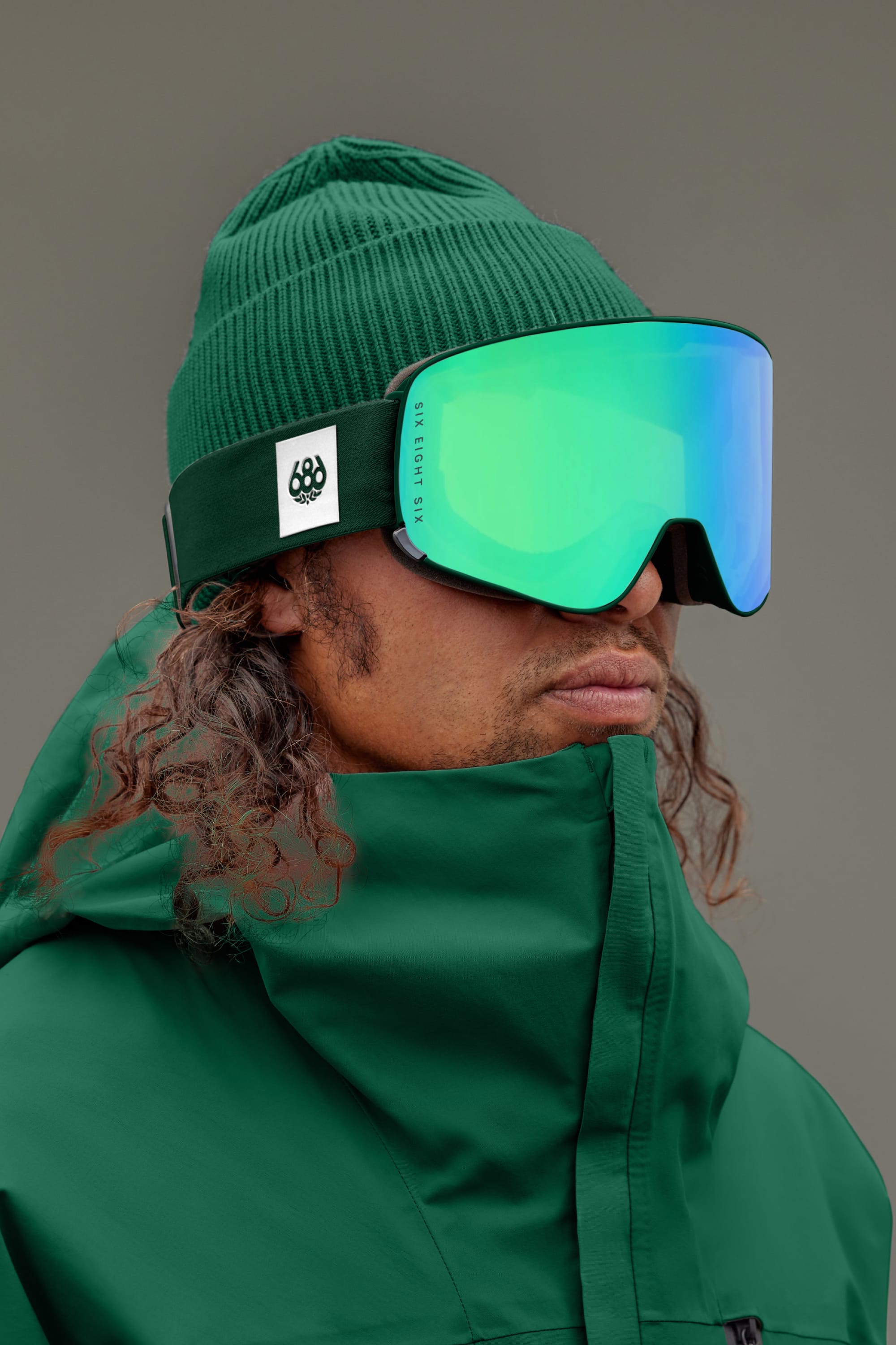 A person wearing a green beanie, a green jacket, and the 686 SMARTY® Magnetic Goggle by 686 stands against a plain background. Their long, curly hair is visible beneath the beanie.